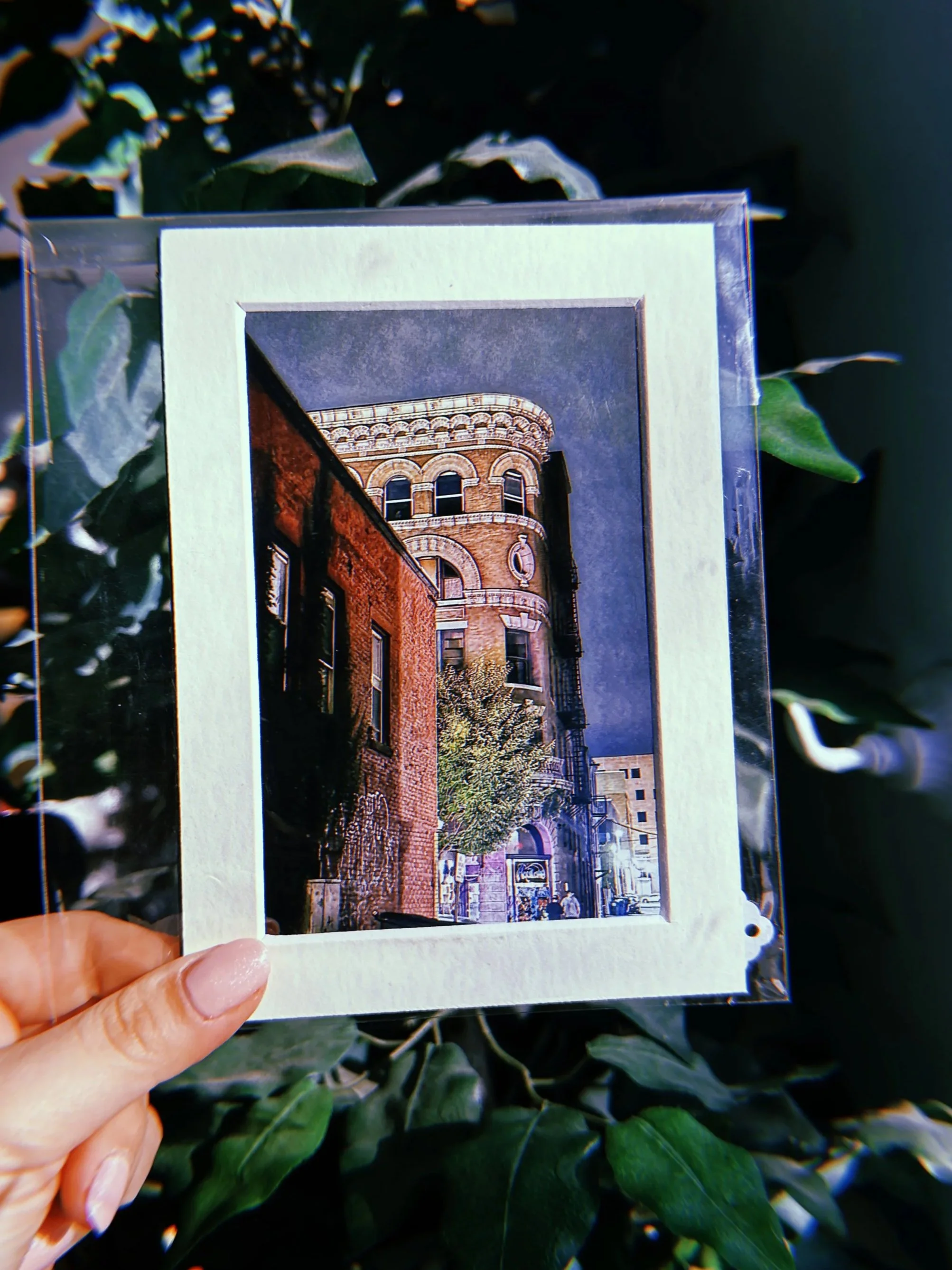A hand holding a framed photograph or postcard of a historic brick building with rounded architectural details and trees in front, against a leafy background.