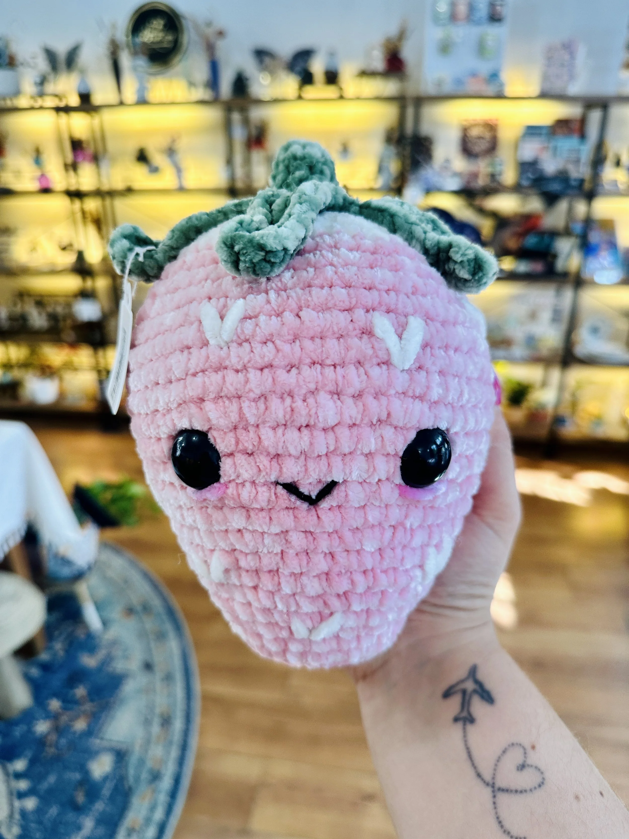 A hand holding a plush stuffed strawberry with a cute face, black eyes, and a small embroidered smile. The strawberry is pink with white seed details and green leaves on top. In the background, there are shelves with various items and a part of a blue rug on a wooden floor.