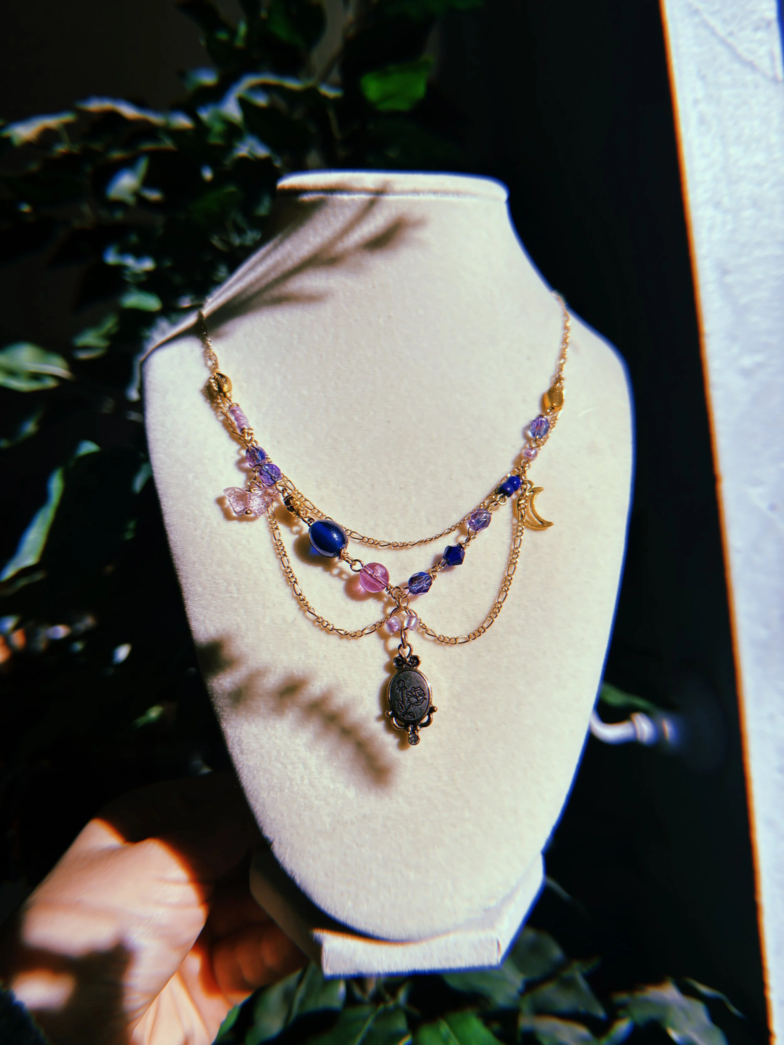 Layered gold necklace with purple, pink, and blue beads and a black pendant with a floral design displayed on a beige jewelry stand.