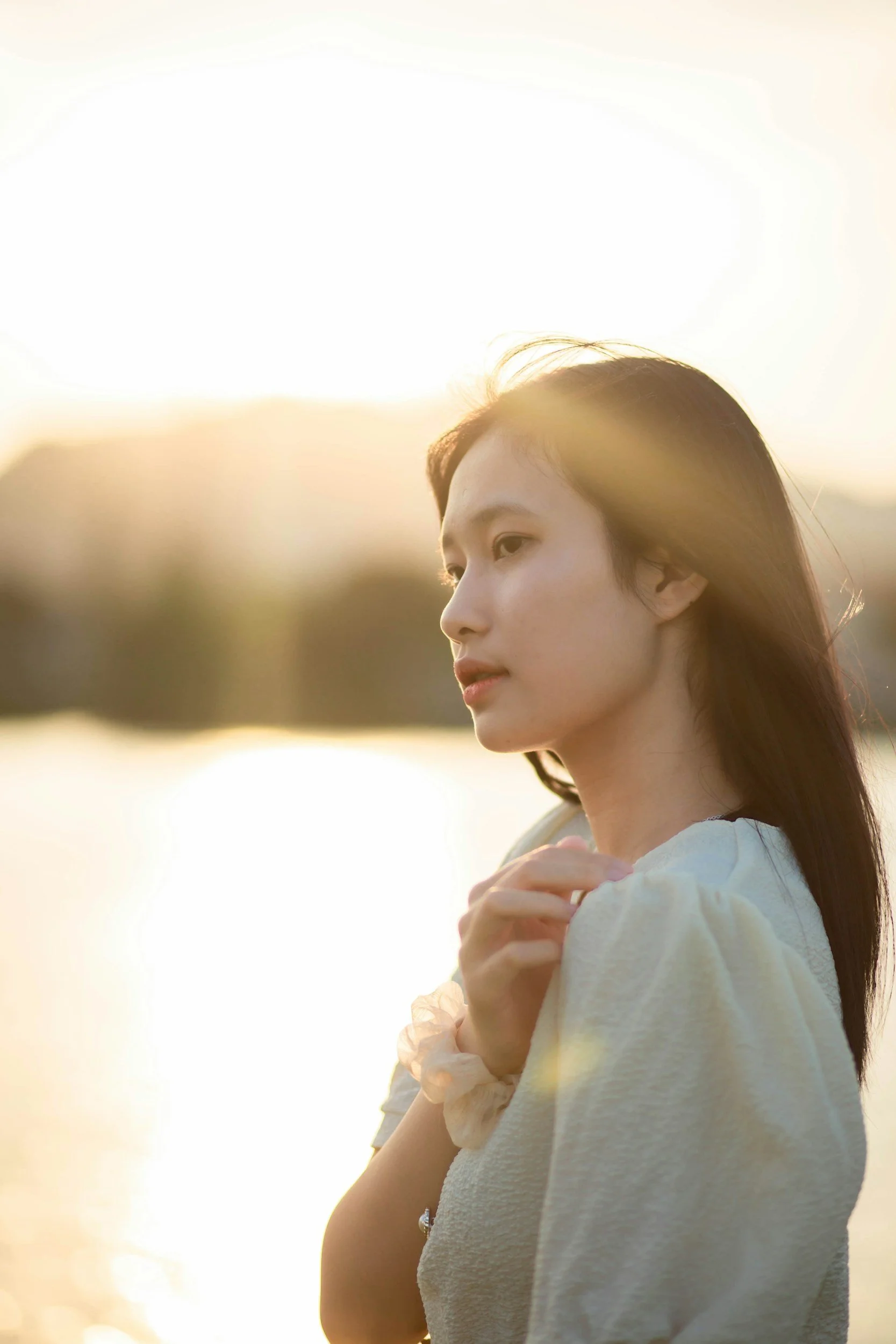 A young woman with long dark hair and light skin is standing outdoors during sunset, wearing a cream-colored top with puffed sleeves. She is holding her hands close to her chest in a peaceful pose.
