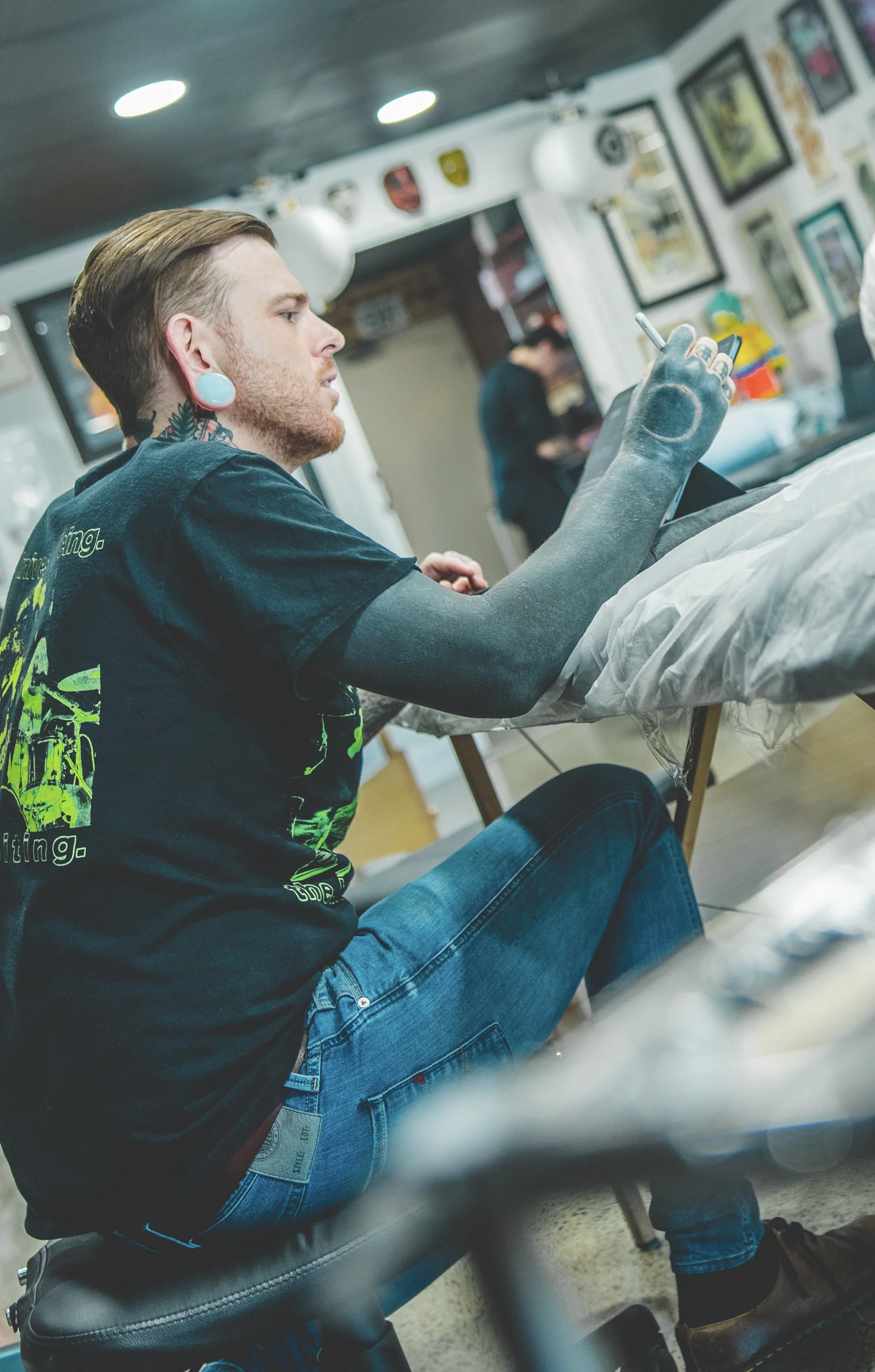 A young man with tattoos, piercings, and colorful earrings is sitting on a tattoo table in a tattoo shop, looking at his phone. The shop has framed artwork and shields hanging on the walls, with a person in the background.