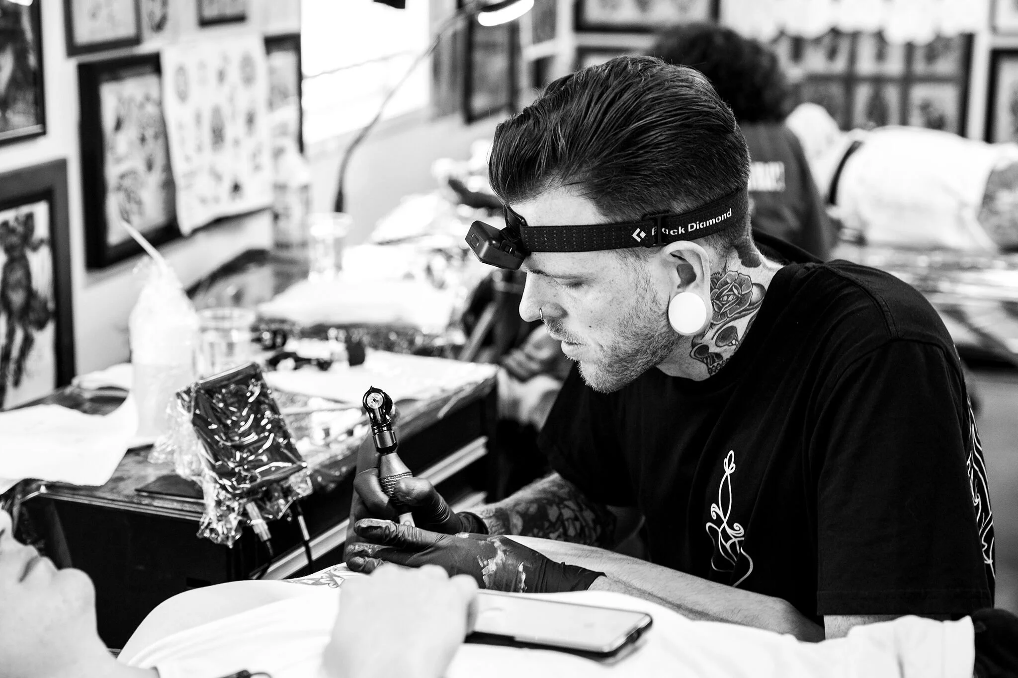 A man with tattooed neck and ears, wearing glasses with a headlamp, permanently inking a tattoo in a tattoo parlor.