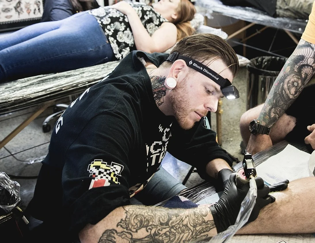 A tattooed man wearing black gloves and a headlamp tattoos a person’s arm in a tattoo parlor, with a woman lying on a table in the background.