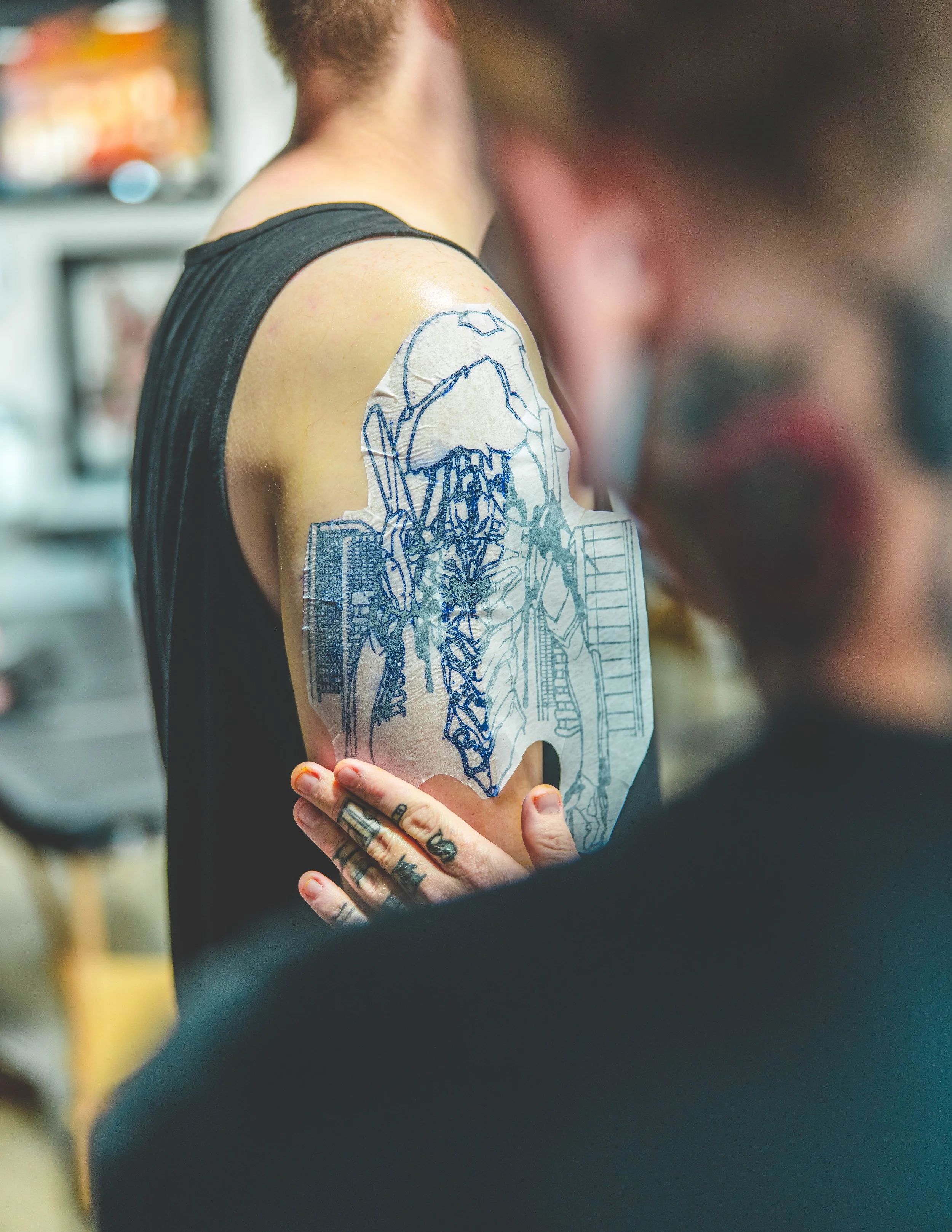 A person with tattoos on their fingers is holding their arm, which has a surgical dressing with a sketch of a mechanical or robotic structure taped to it. The person's head is blurred in the foreground.