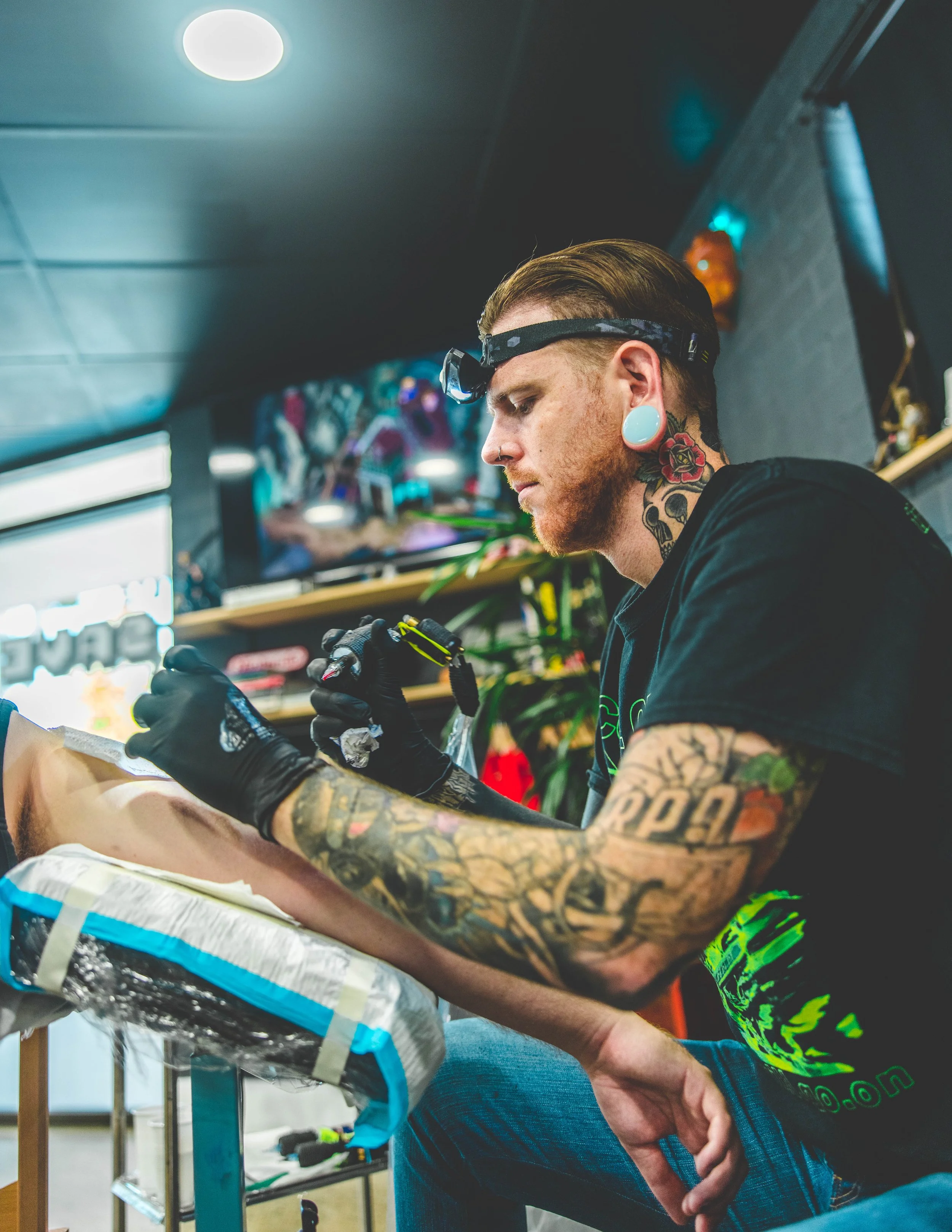 A tattooed man with gauges in his ears and a headlamp is tattooing a person's leg in a tattoo shop, with shelves and artwork in the background.