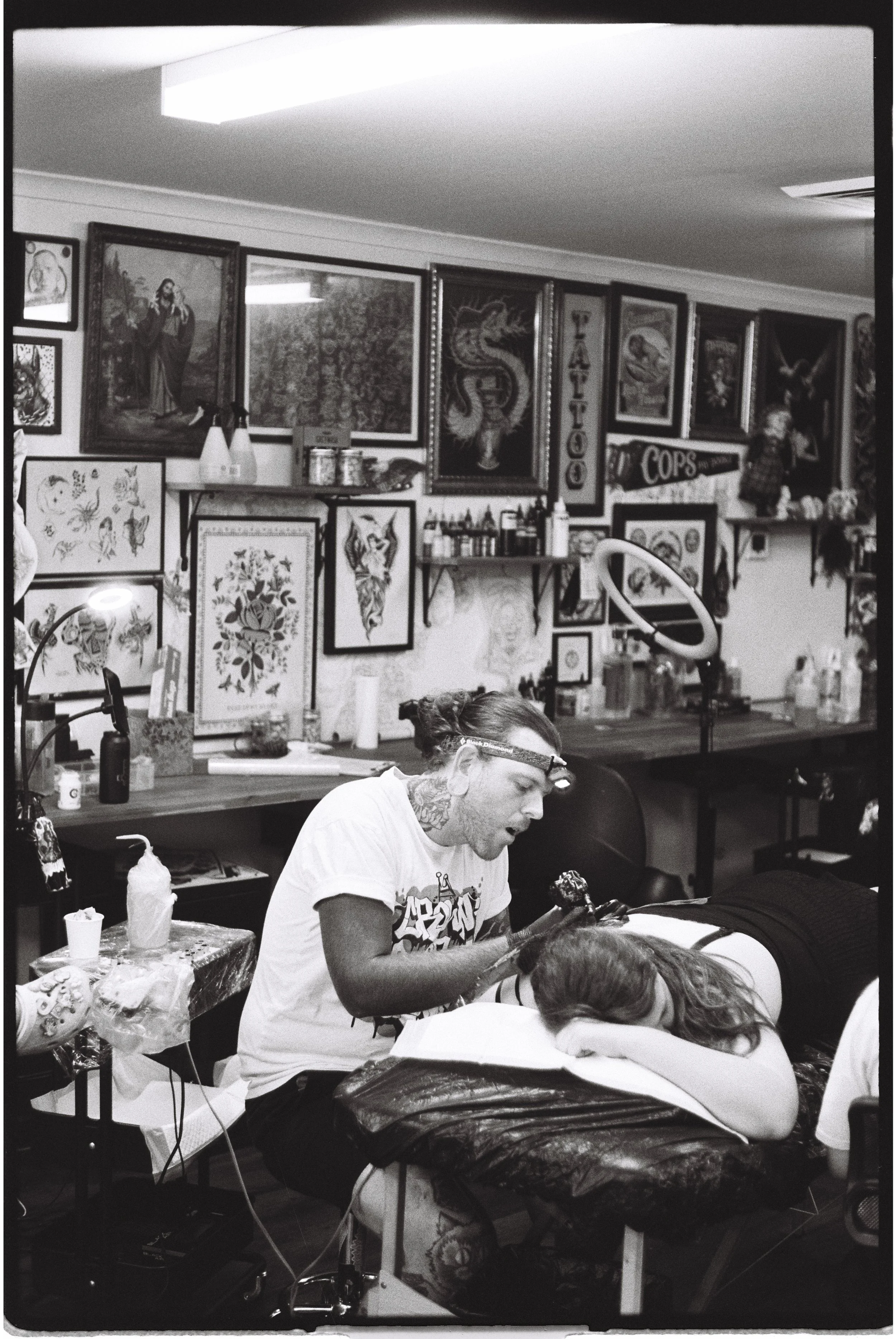 A tattoo artist applying a tattoo to a client lying on a tattoo bed, in a tattoo parlor decorated with various framed artwork, including traditional and tattoo-themed images.