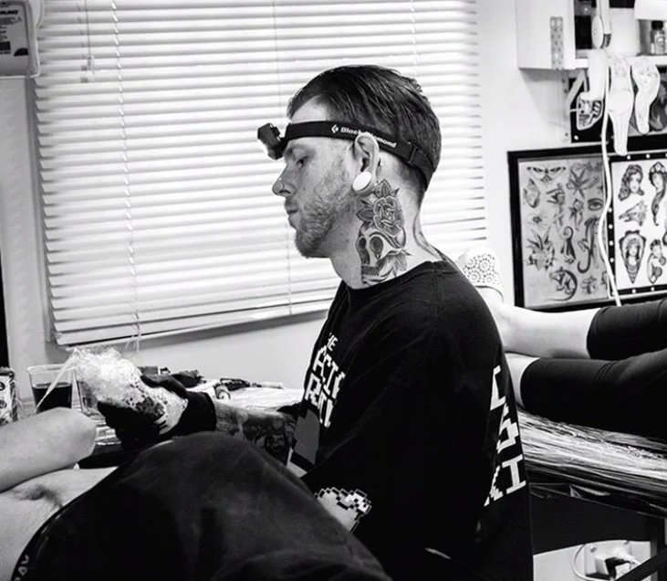 Tattooed man with headphones and ear plugs sitting in a tattoo shop and getting a tattoo on his hand.