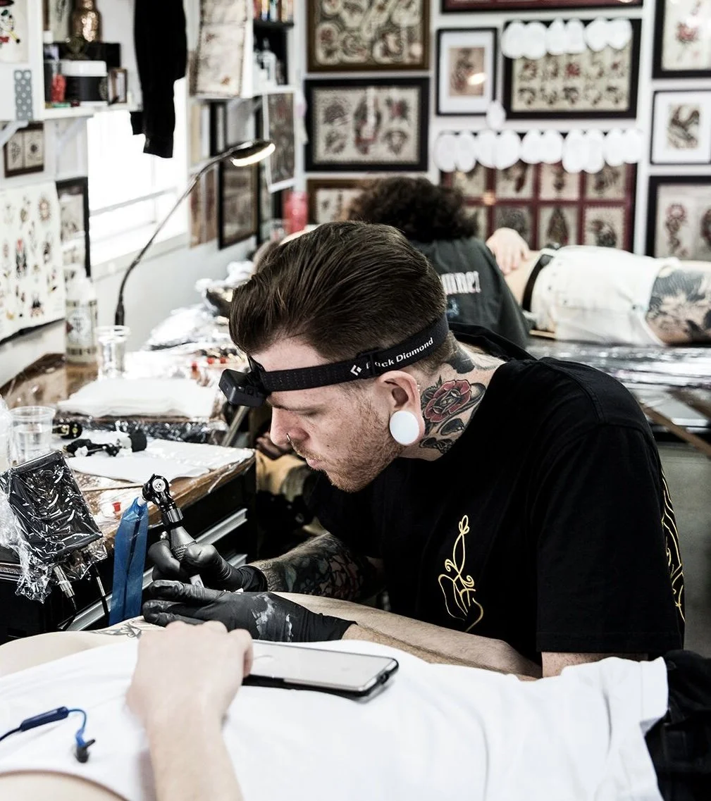 A tattoo artist with tattoos, gauged ears, and a headlamp tattooing a person in a tattoo studio with framed artwork on the walls.
