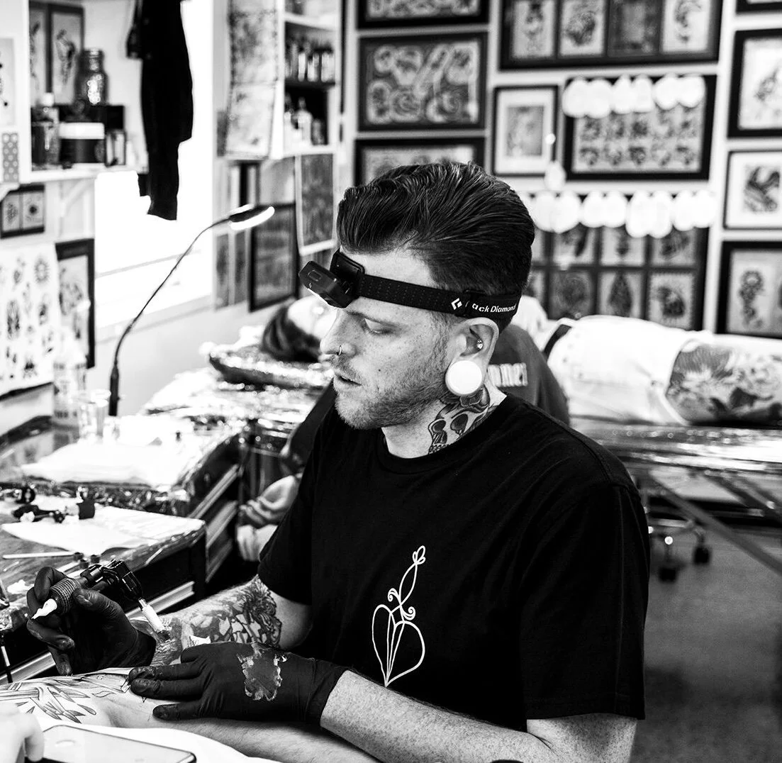 A man with tattoos, a nose ring, and large gauge earrings tattooing someone's arm in an art studio with tattoo designs on the wall.