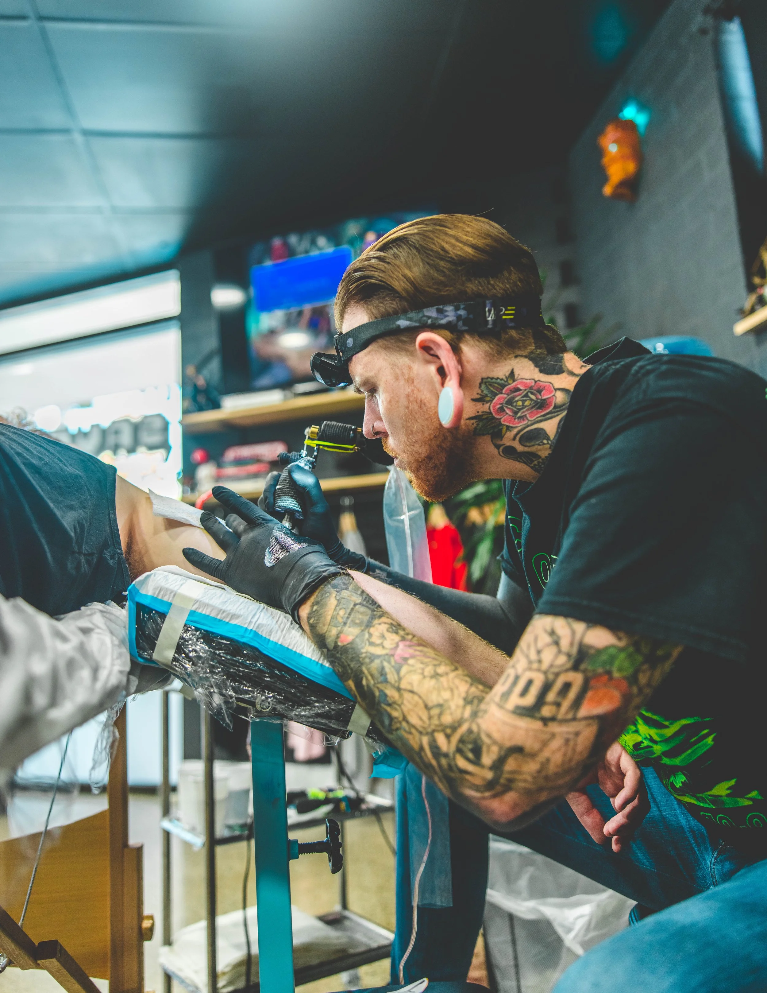 Tattooed man wearing glasses tattooing a person's arm while working in a tattoo shop with various supplies and equipment visible in the background.
