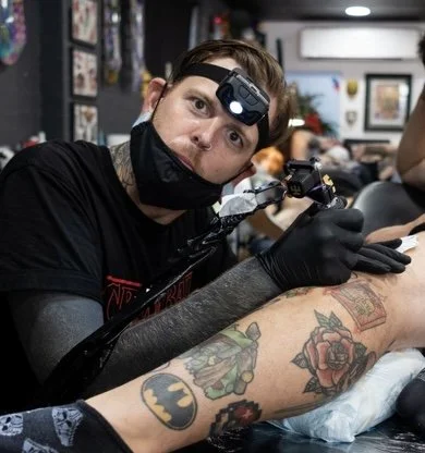 Tattoo artist working on a client’s arm in a tattoo shop, with equipment and artwork on the walls visible in the background.