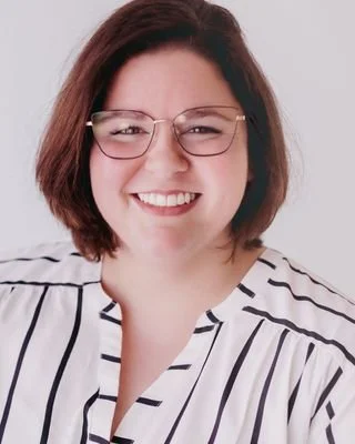 A woman with shoulder-length brown hair, wearing glasses with a thin gold frame and a white blouse with black horizontal stripes, smiling at the camera.