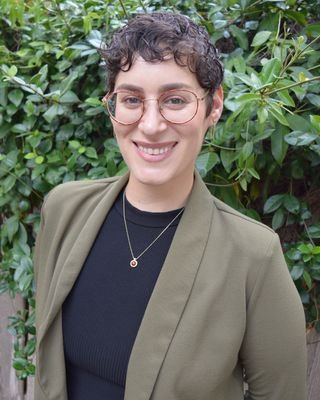 A woman with short curly hair and glasses wearing a black shirt, an olive green blazer, and a necklace, smiling in front of green foliage.