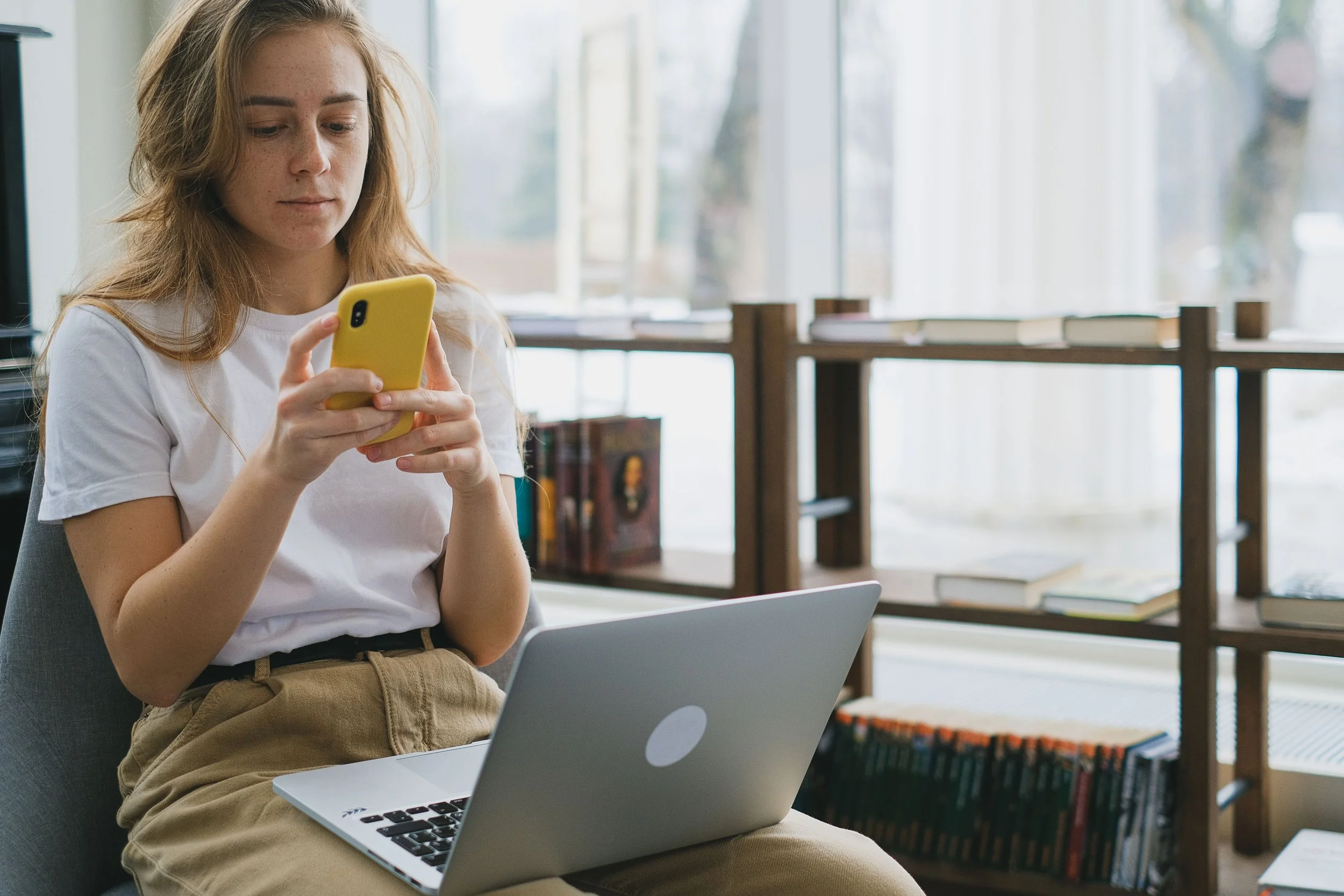 Focused woman scrolling mindlessly on her yellow smartphone while sitting in front of an open laptop.