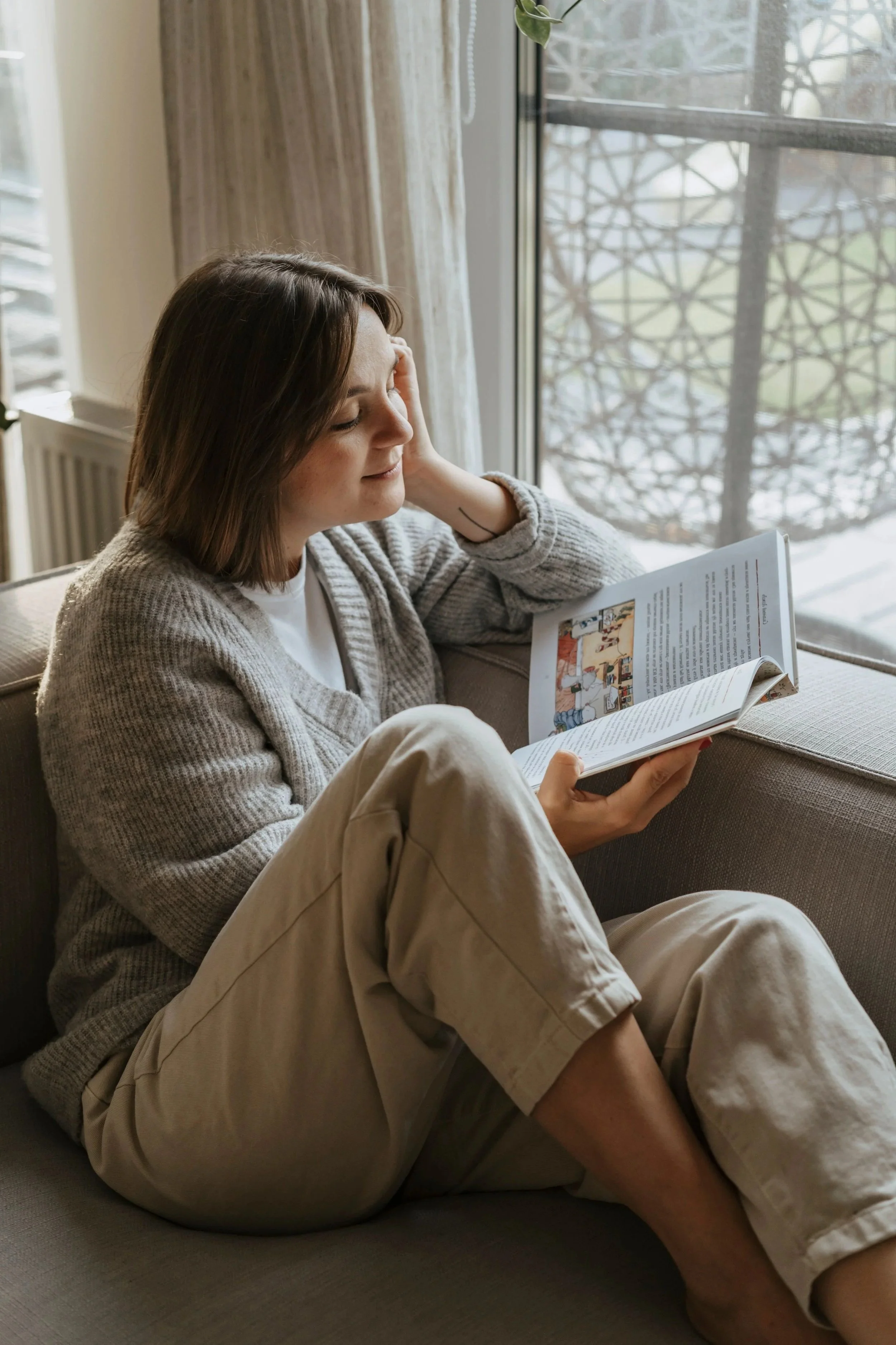 A woman with brown hair is sitting on a gray couch, reading a magazine with colorful illustrations. She is wearing a beige cardigan and beige pants, with her left hand resting on her forehead. Behind her is a window with a metal lattice, and a sheer curtain partially covers the window. The scene appears to be by a sunny window during the daytime.