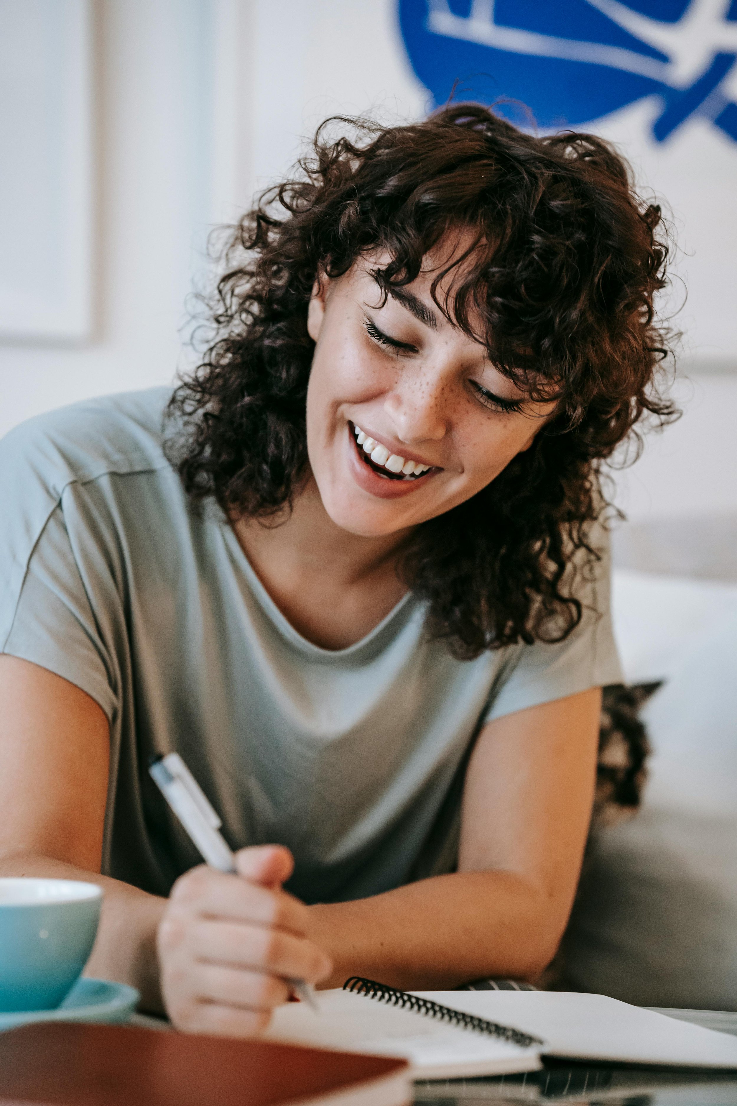 A young woman with curly dark hair smiling while writing in a notebook, with a pen in her right hand, and a cup of tea or coffee nearby.