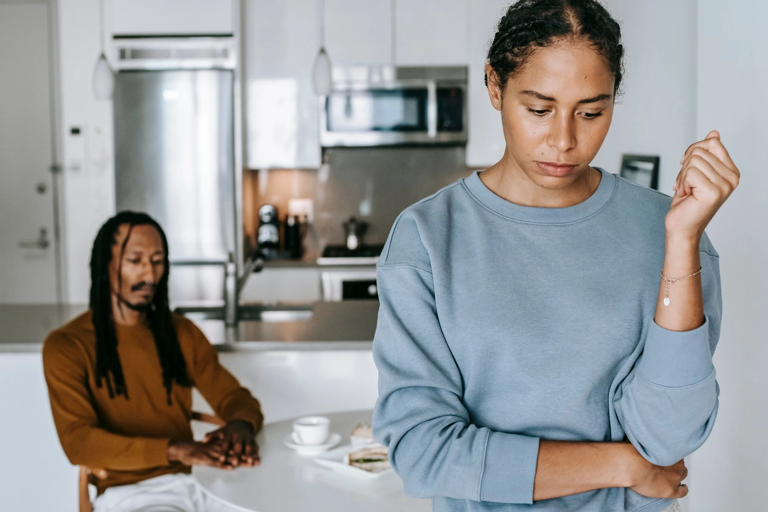 A woman in a blue sweatshirt with curly black hair looks down and appears upset or thoughtful, while another woman with long dreadlocks, wearing a brown top, sits at a kitchen table with her eyes closed, possibly praying or meditating.