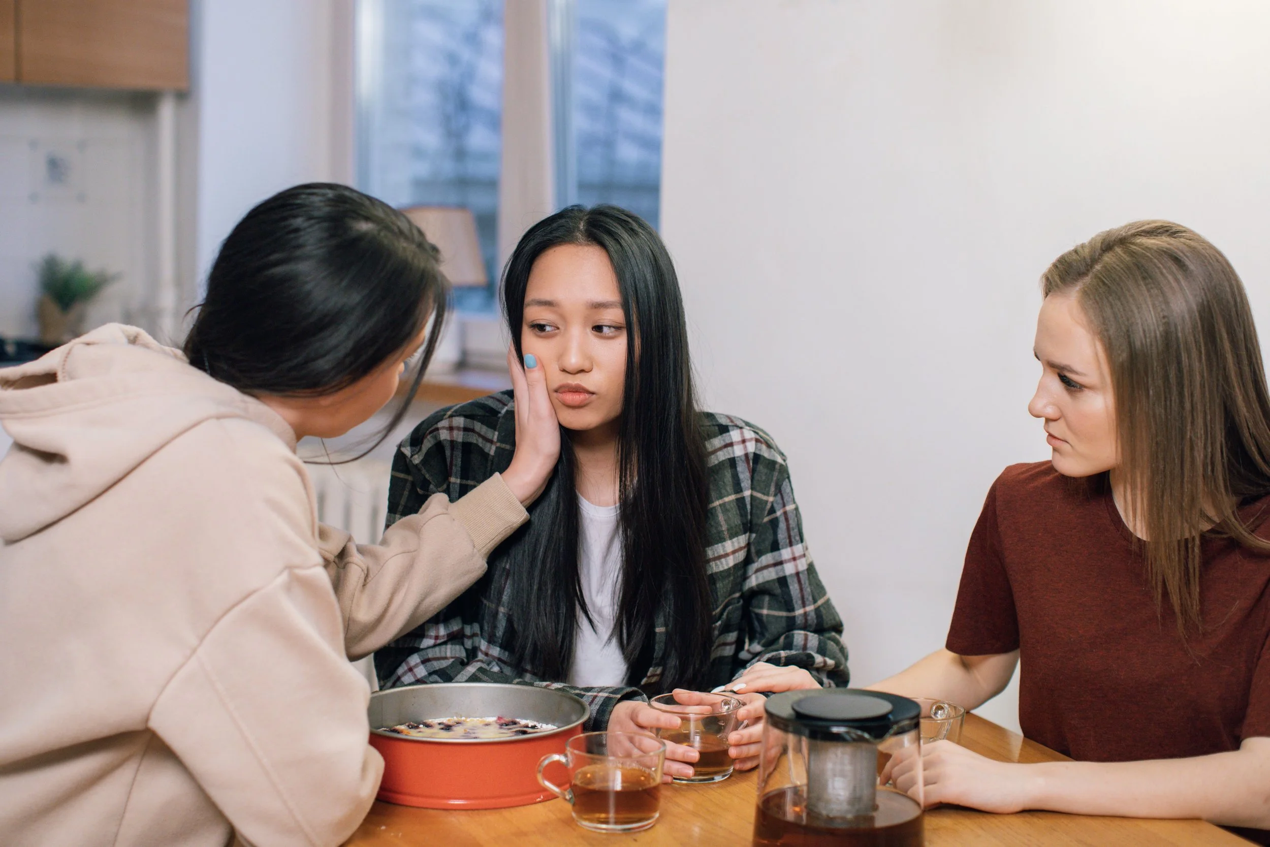 Three women sitting at a table, one woman gently touching another woman's face.