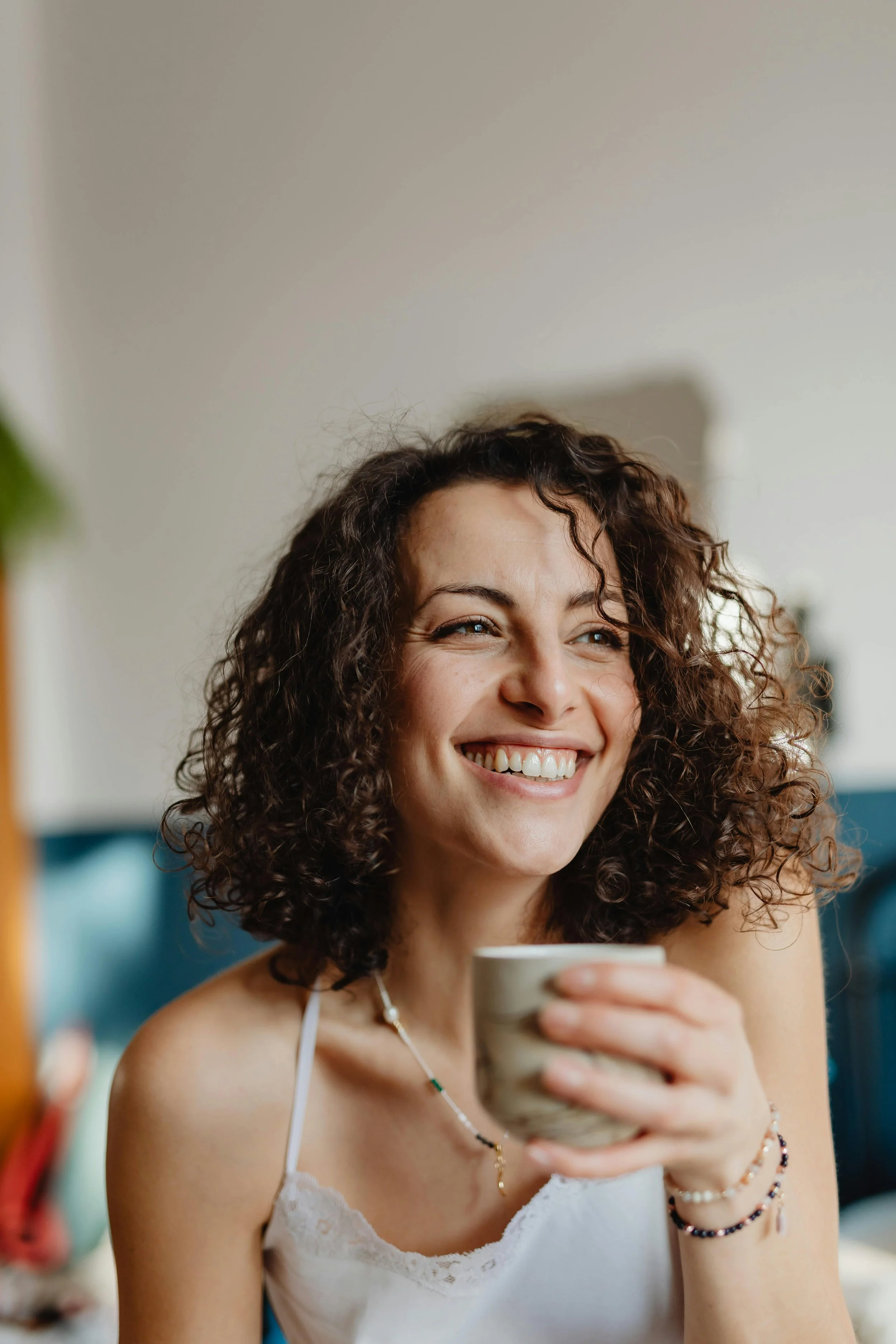 A smiling woman with curly brown hair holding a cup, wearing a white spaghetti strap top and jewelry, sitting indoors.