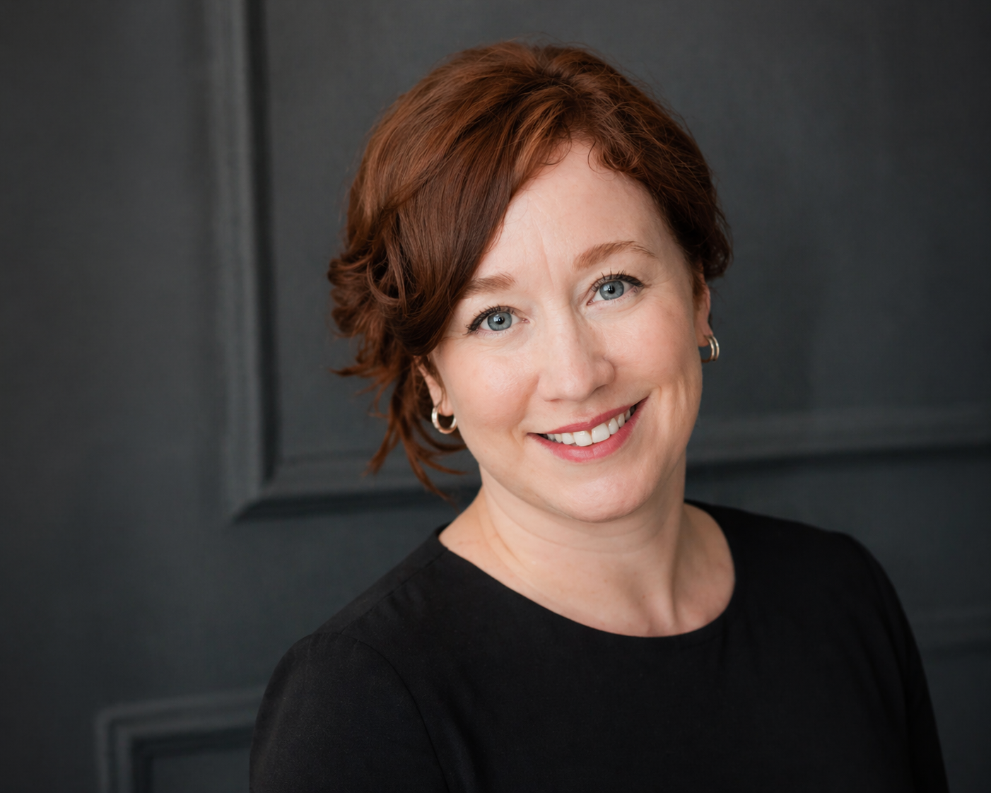 A woman with short red hair and blue eyes smiling, wearing a black top and silver hoop earrings, standing against a dark gray background.