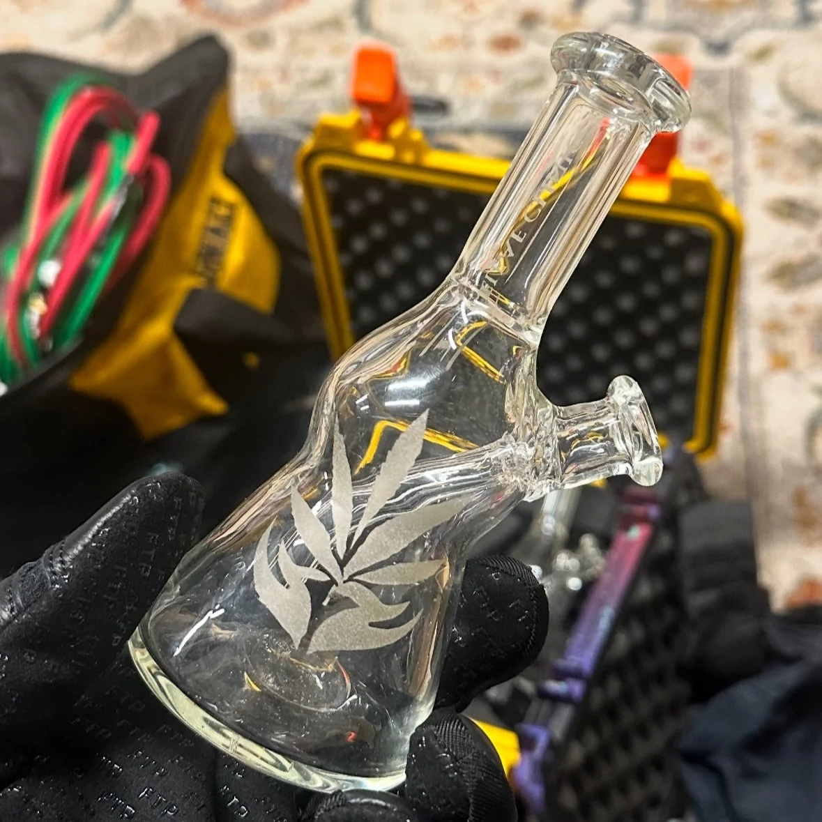 A glass piece in the shape of a small bottle rig with a cannabis leaf design etched on it, being held by a gloved hand, with a black and yellow case and a bicycle in the background.