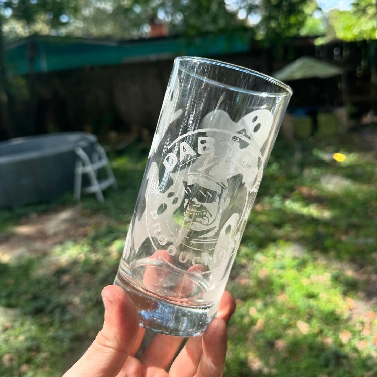 Clear glass with a skeleton wearing sunglasses, surrounded by ghosts, with text 'DAB' and 'RODUC' etched on it, being held outdoors in a grassy backyard.