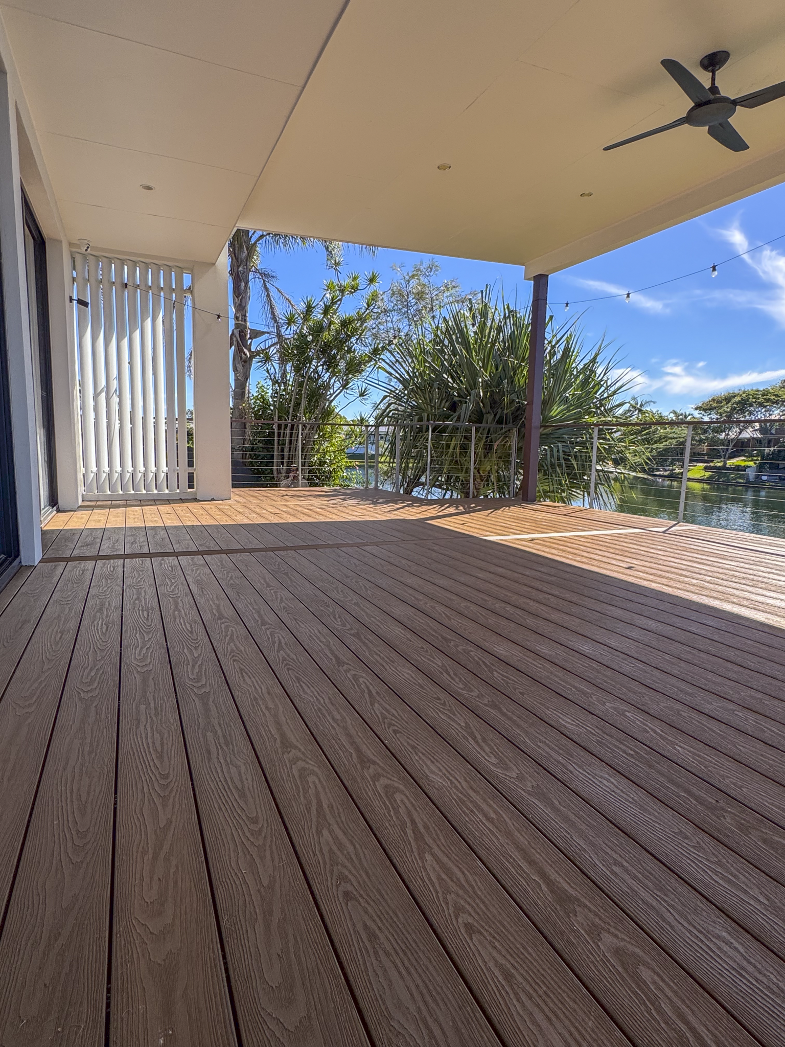 View of a covered outdoor patio with wooden decking, a ceiling fan, string lights, and lush green tropical plants and trees in the background, under a bright blue sky with some clouds.