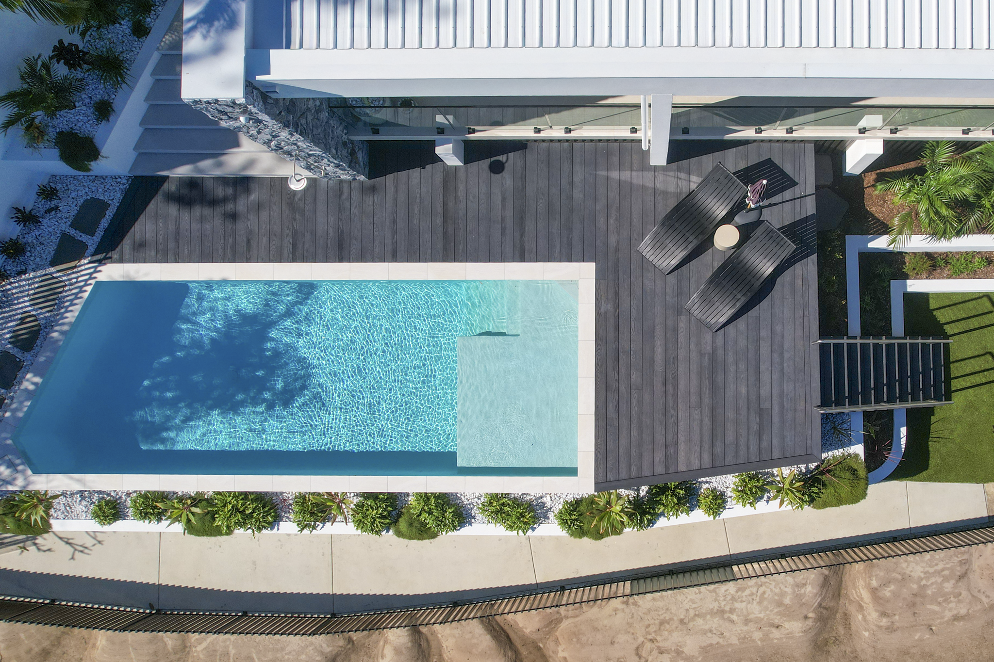 An aerial view of a modern house with a swimming pool, wooden deck, and surrounding landscaping with plants and trees.
