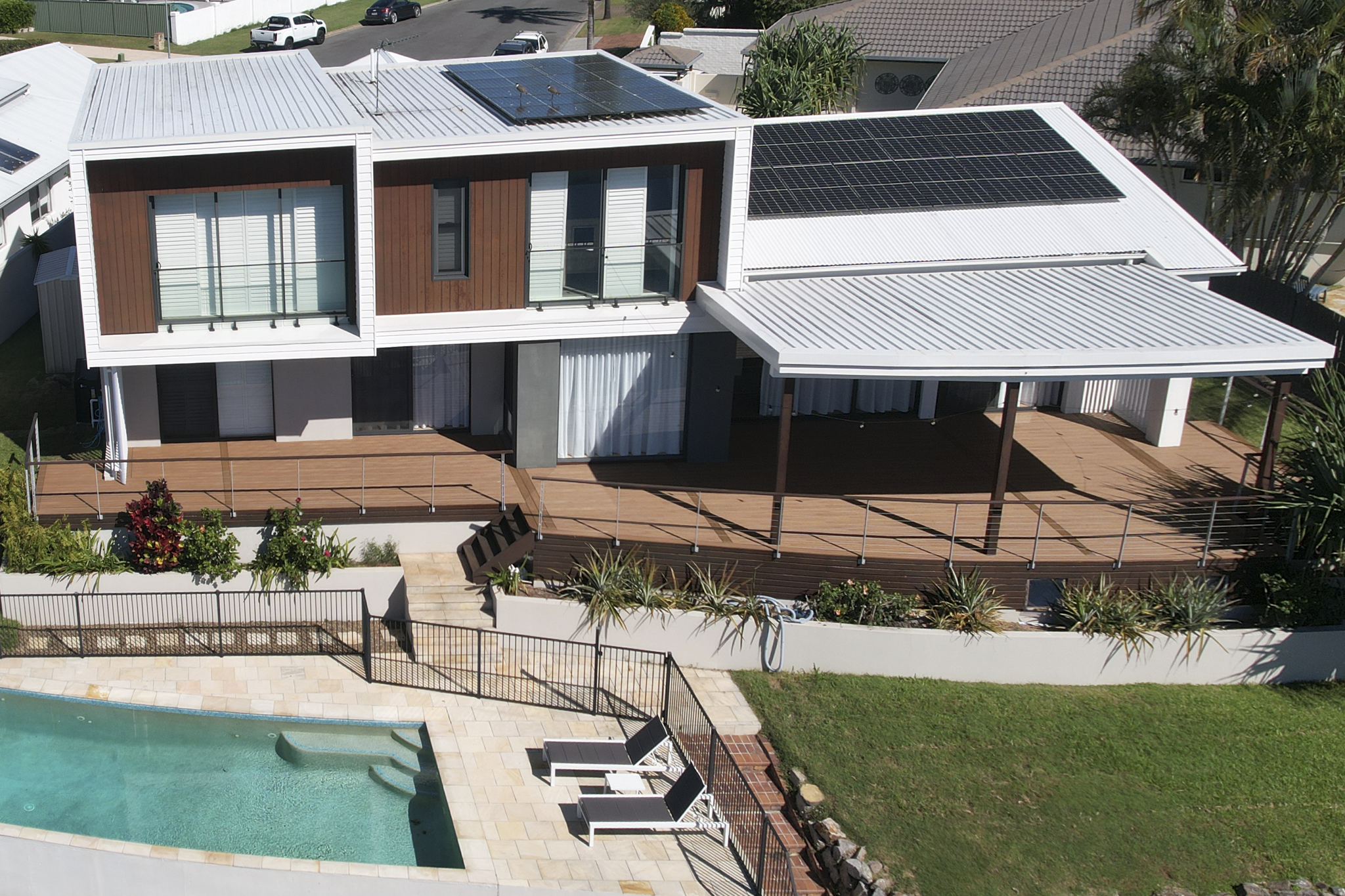 A modern two-story house with a backyard featuring a swimming pool, a wooden deck with lounge chairs, and a metal roof with solar panels. The house has large windows and a balcony on the upper floor.