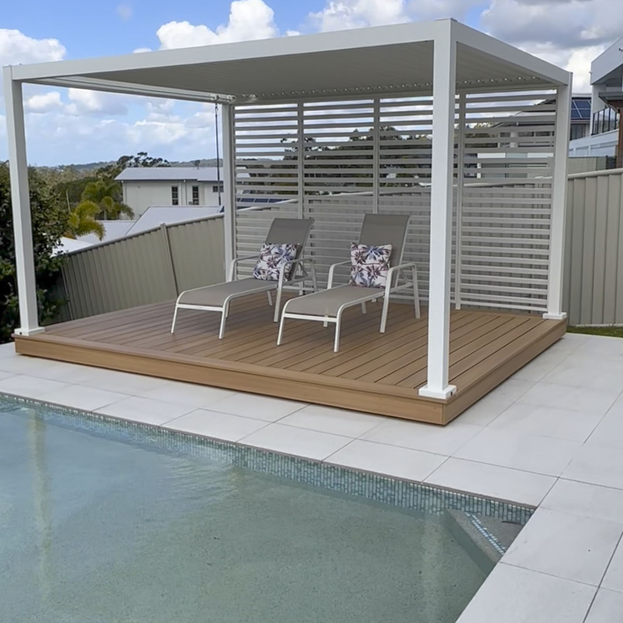 View of an outdoor swimming pool with a wooden deck and lounge chairs. A white pergola covers part of the deck, with cloudy skies overhead.