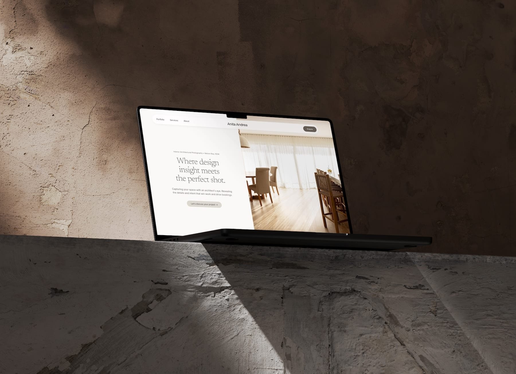 An open Macbook sitting on a concrete shelf showing a half and half split homepage design with a floating nav bar and elegant typography, the homepage of Anita Andrea Photography, an interior photographer in Adelaide