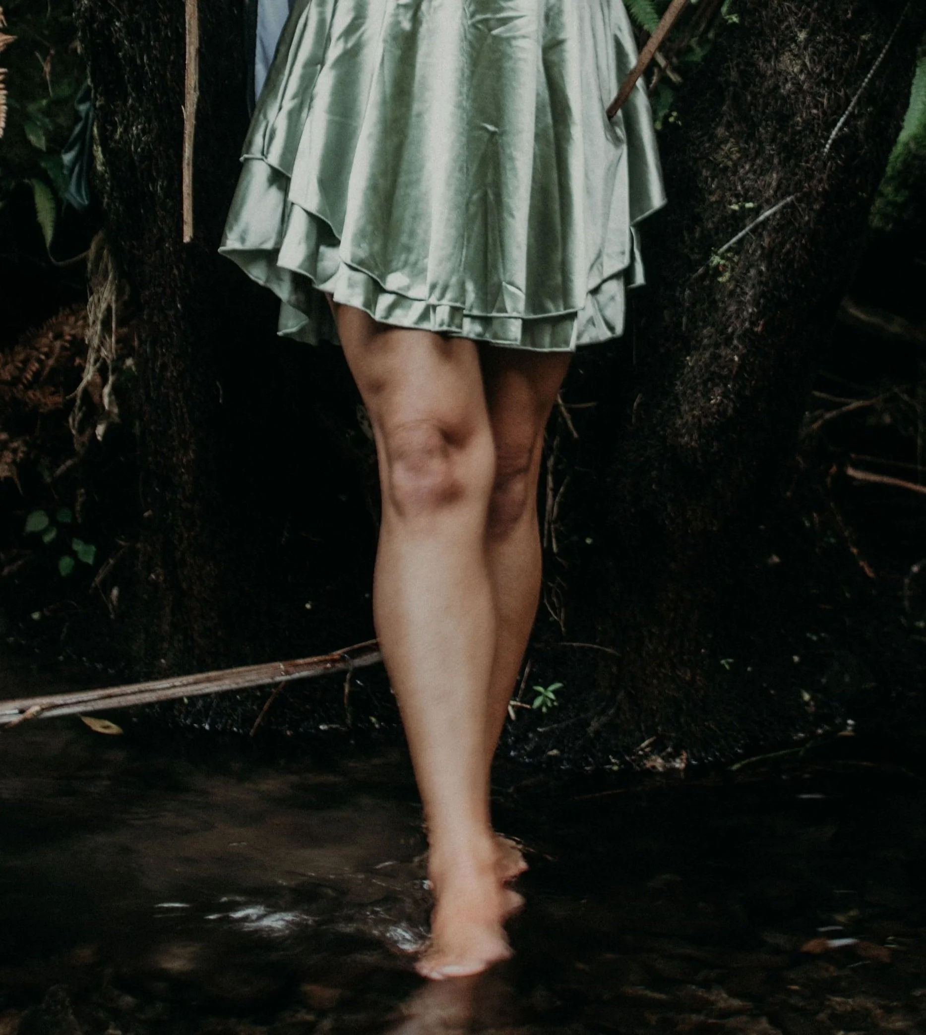 Pierna de una mujer con falda de satén verde y pies descalzos en agua, rodeada de árboles y vegetación.