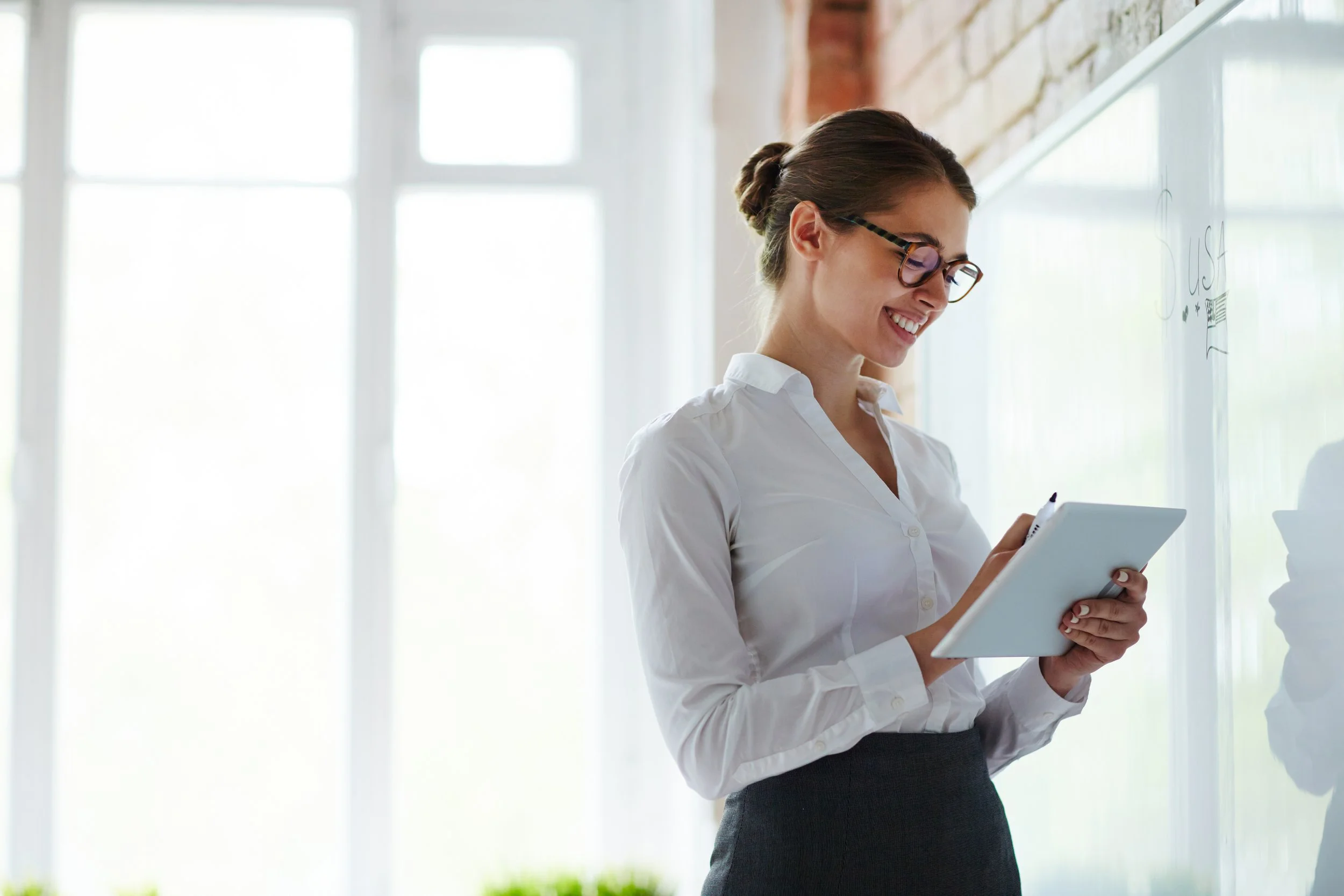 A woman wearing glasses, a white shirt, and a dark skirt, smiling while looking at a tablet in her hands near a whiteboard in a bright room with large windows.