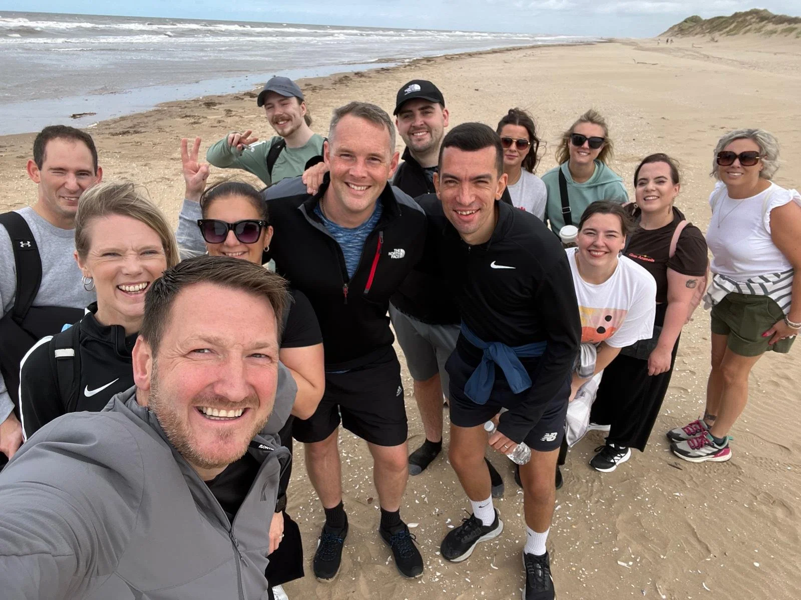Group of friends smiling for a selfie on a sandy beach with ocean waves and cloudy sky in the background.