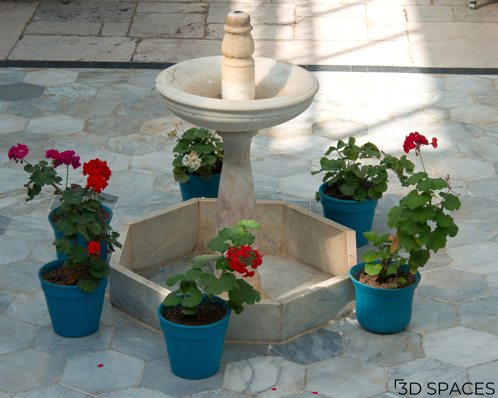 Small marble water fountain centerpiece surrounded by potted red and white geraniums on a stone patio courtyard.