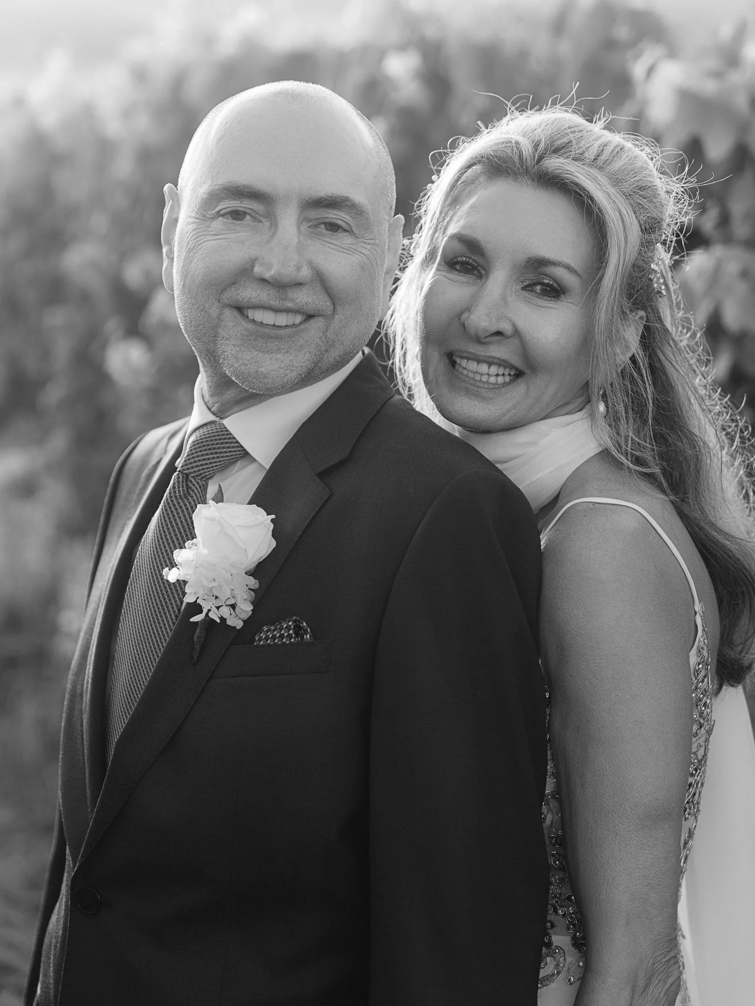 A happy couple, a man in a suit with a boutonniere and a woman in an elegant dress, smiling outdoors.