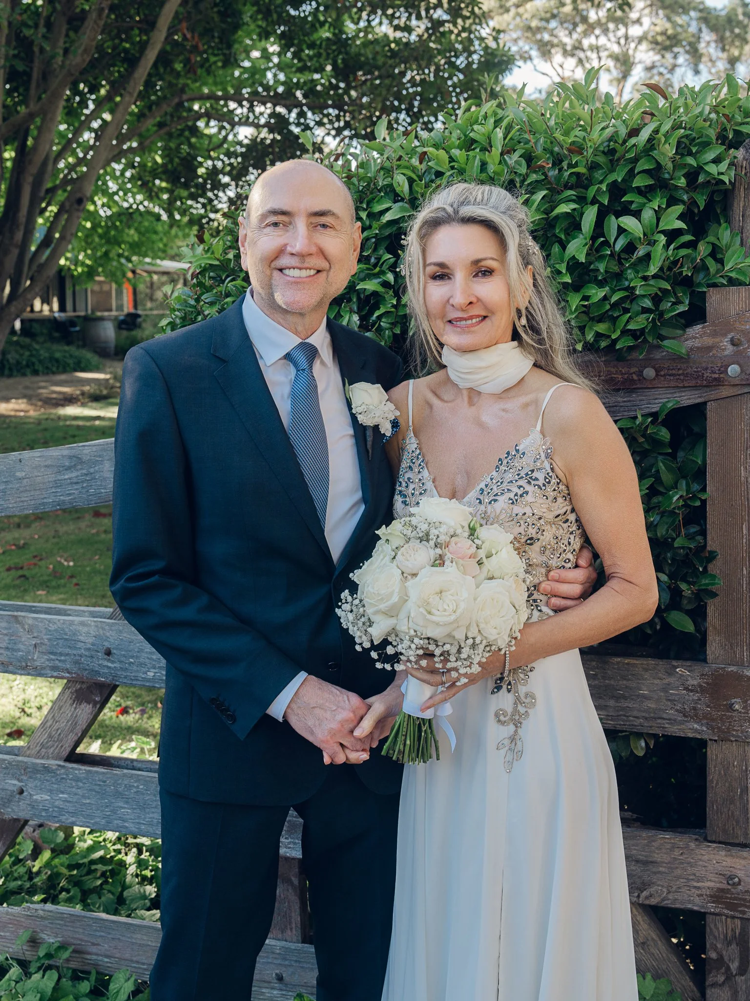 A man and a woman standing outdoors, dressed in formal attire, smiling, with the man in a suit and the woman in a white gown holding a bouquet of white roses, greenery, and baby's breath, in front of a wooden fence and green foliage.