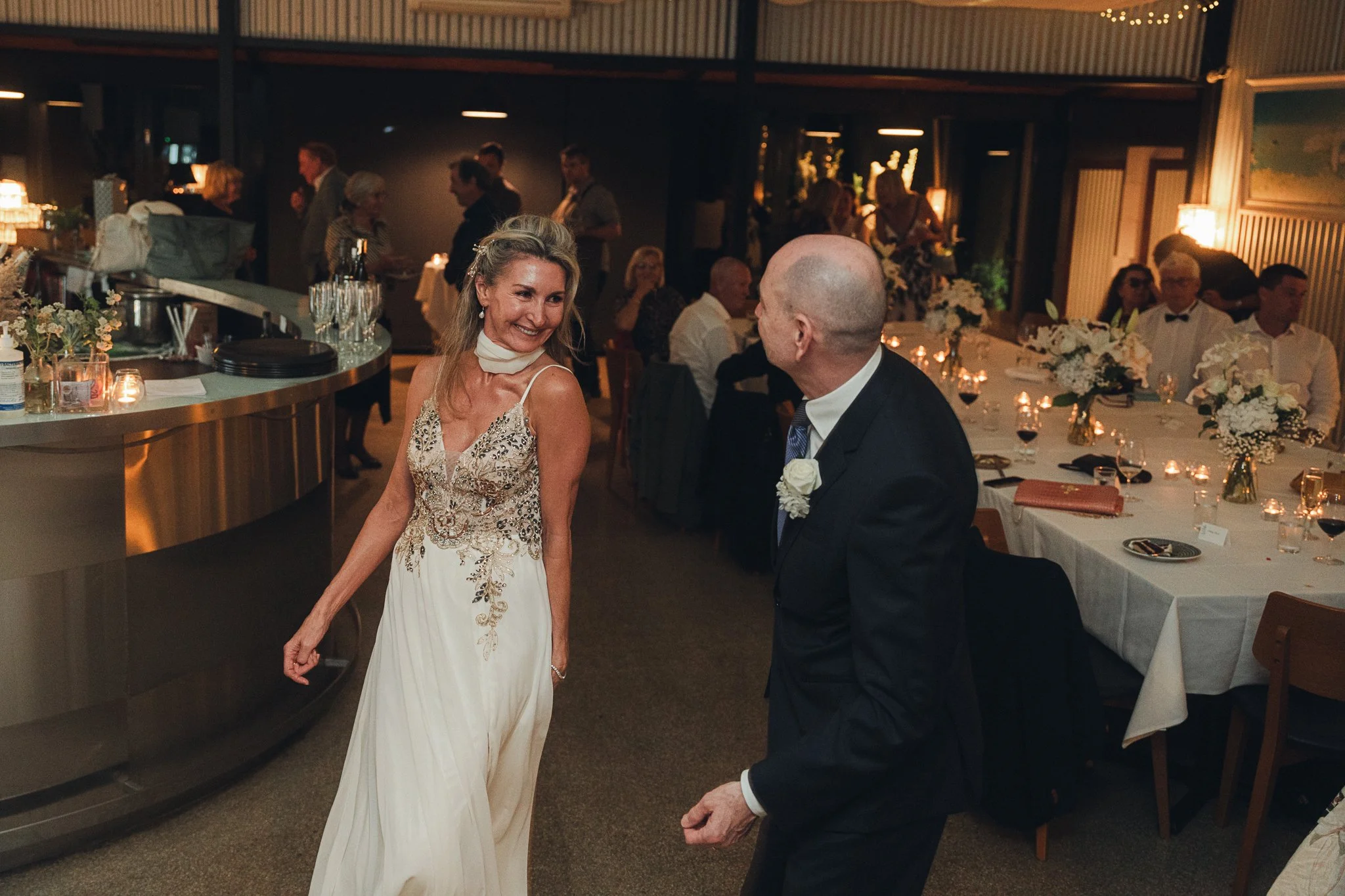 A woman in a white and gold embellished dress dancing with a bald man in a black suit at a wedding reception with guests seated at decorated tables in the background.