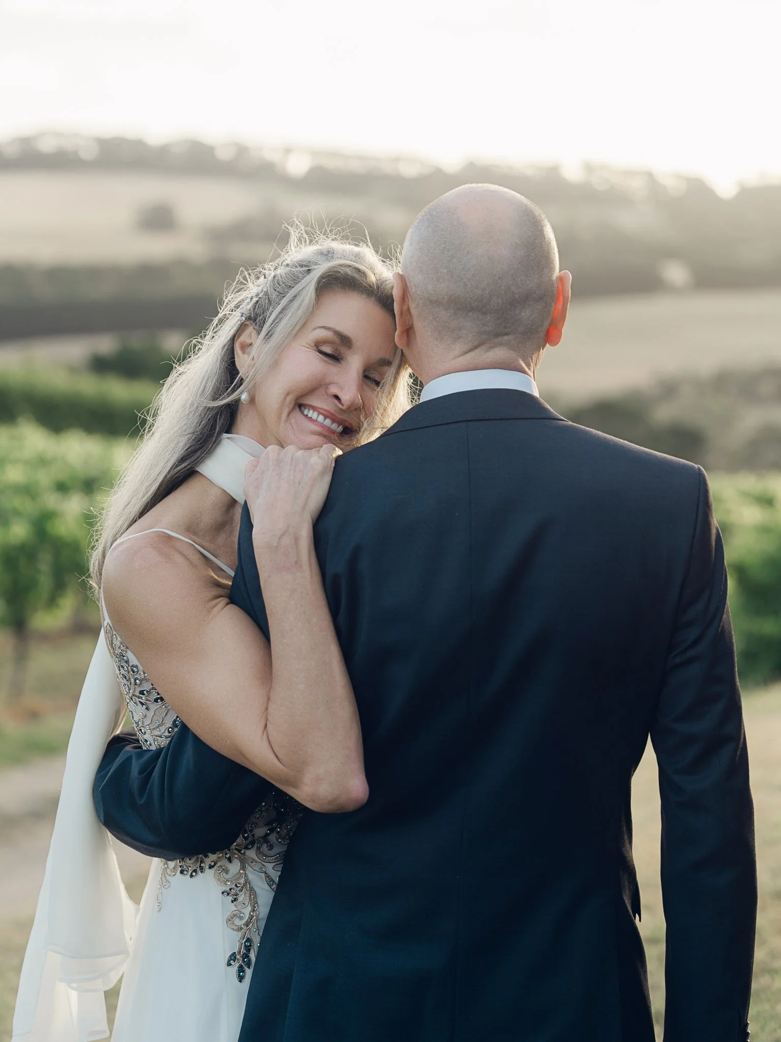 Hobart wedding photographer capturing A smiling woman with long blonde hair embraces a man in a black suit outdoors. They are standing in a green field with hills in the background, during sunset.