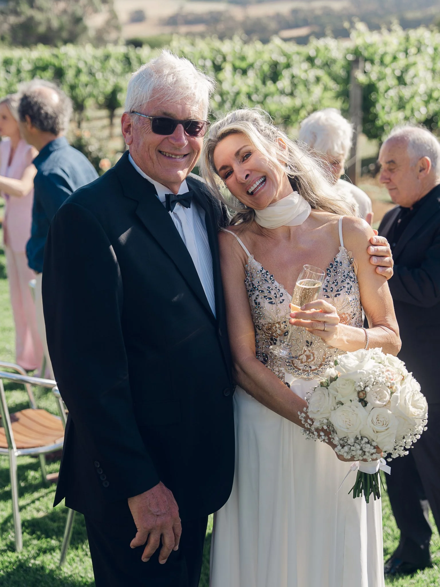 A joyful wedding scene with an older man in a black tuxedo and woman in a wedding dress, holding a bouquet of white roses, outdoors in a vineyard, smiling and celebrating.