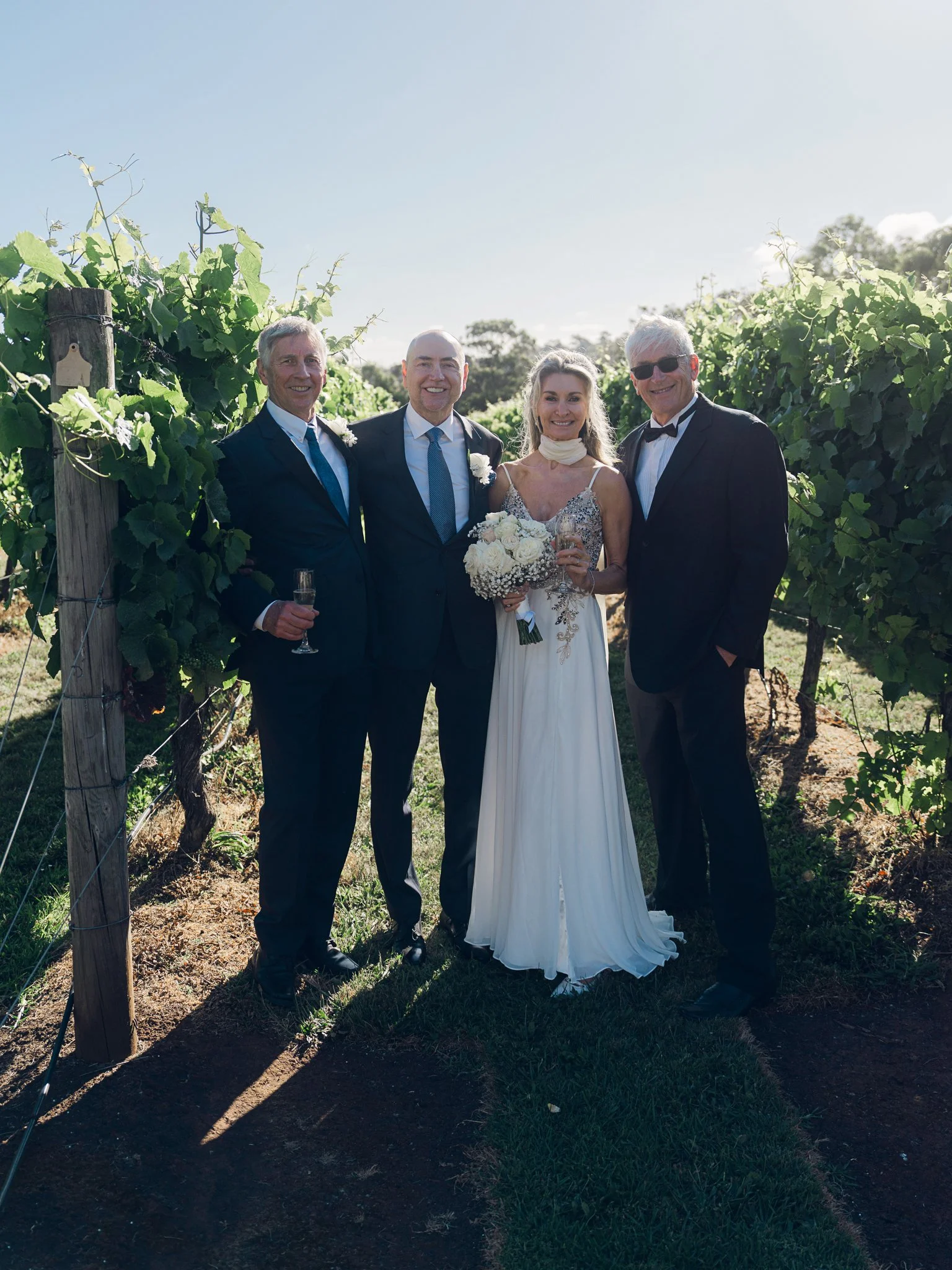 Wedding party of four people in a vineyard, with the bride in a white dress holding a bouquet, and the groom and two men in suits, celebrating outdoors on a sunny day.