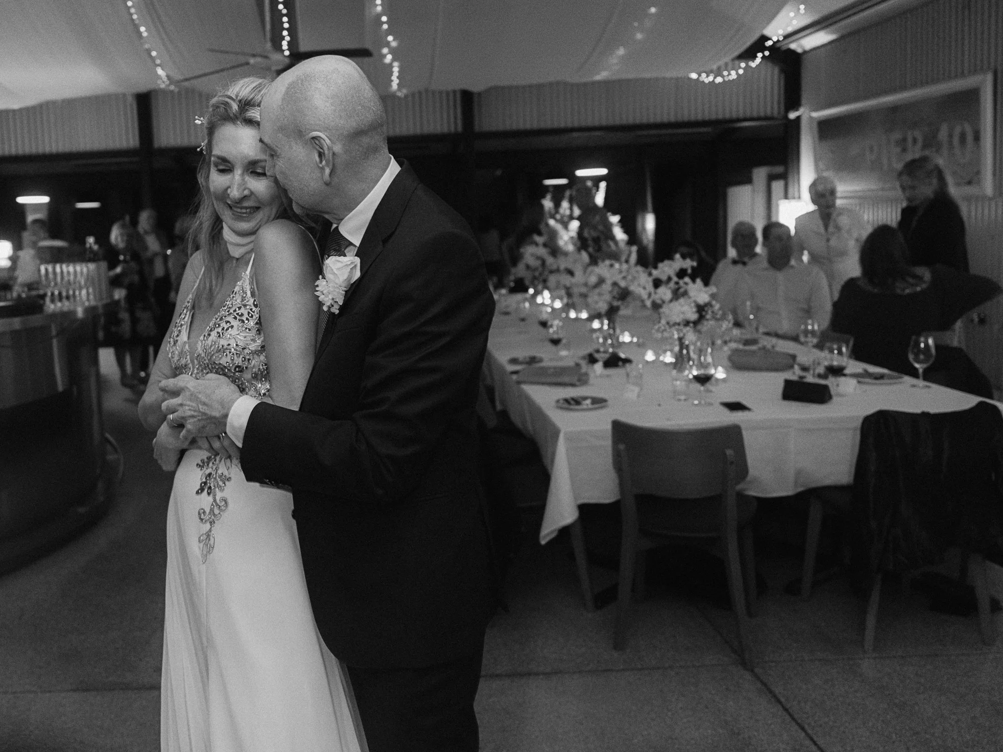 A bride and groom dancing closely at a wedding reception, surrounded by guests seated at decorated tables with flowers and wine glasses.