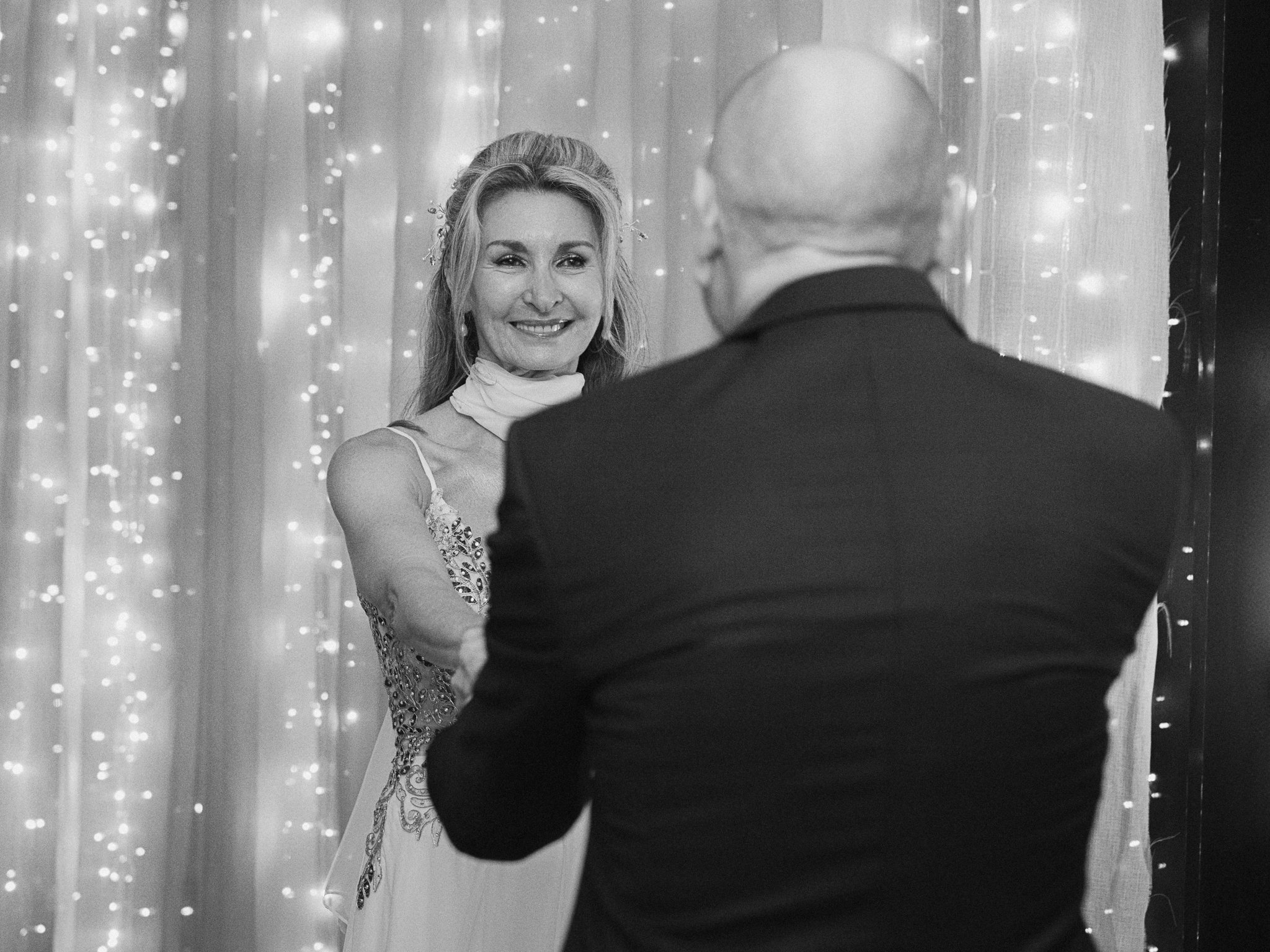 A woman in a wedding dress smiling at a man in a tuxedo during a wedding ceremony, with curtains and twinkling lights in the background.