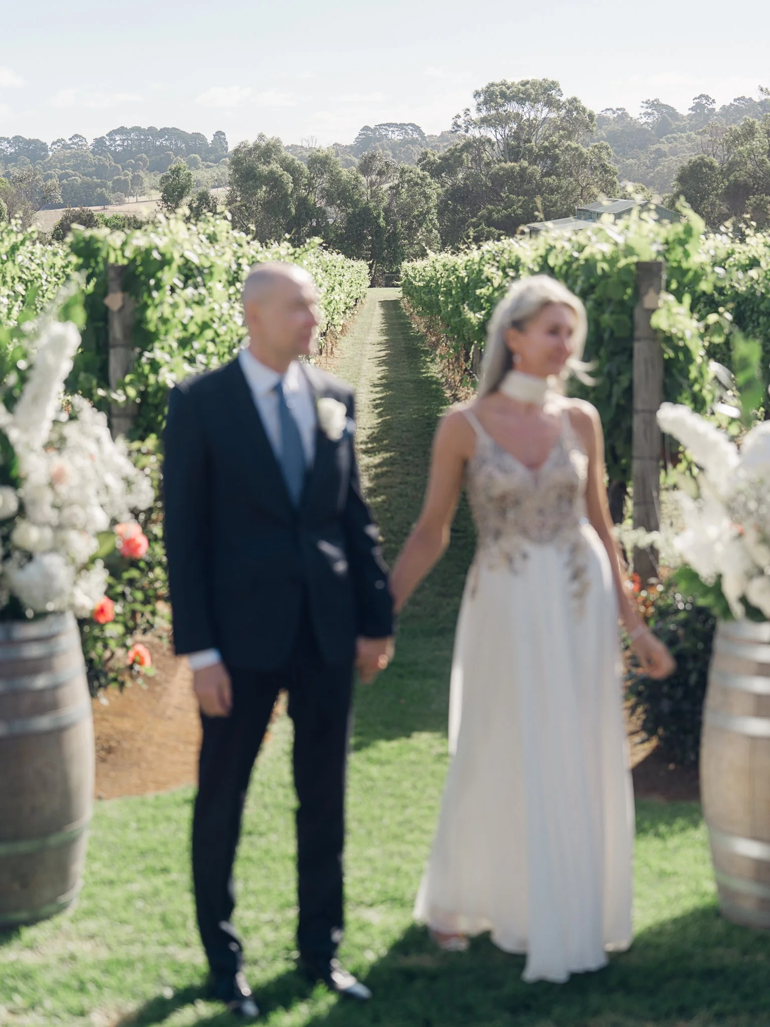 A couple dressed in wedding attire standing hand in hand in a vineyard, surrounded by flowers and greenery on a sunny day.