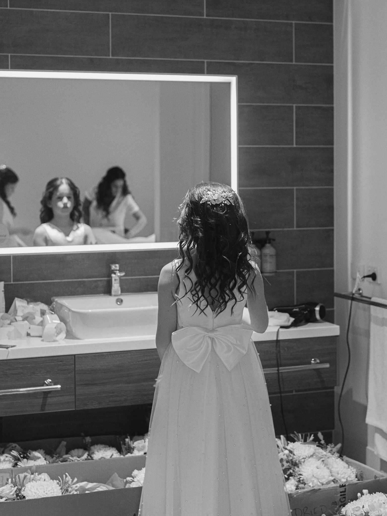 Hobart wedding photographer capturing A young girl in a white dress with a large bow on the back stands in front of a mirror, looking at her reflection in a cake-filled room, possibly preparing for a special occasion.