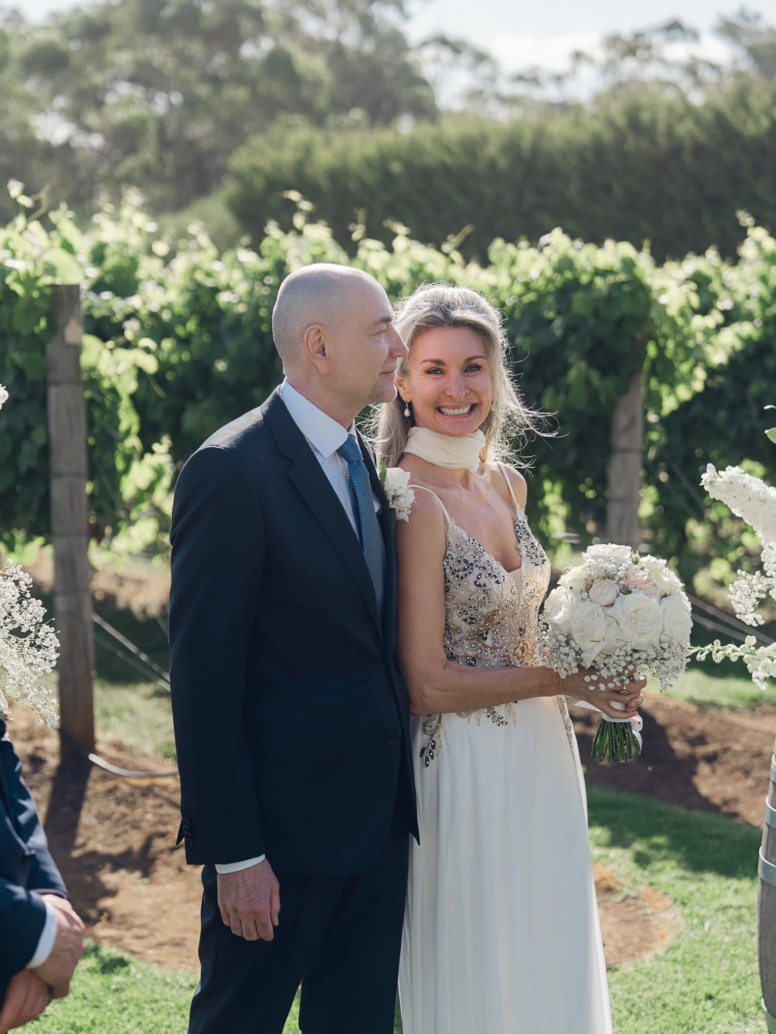 A bride and groom standing together outdoors, with the bride smiling brightly and holding a bouquet of white flowers, while the groom stands beside her in a dark suit. The background features lush green vineyard rows.