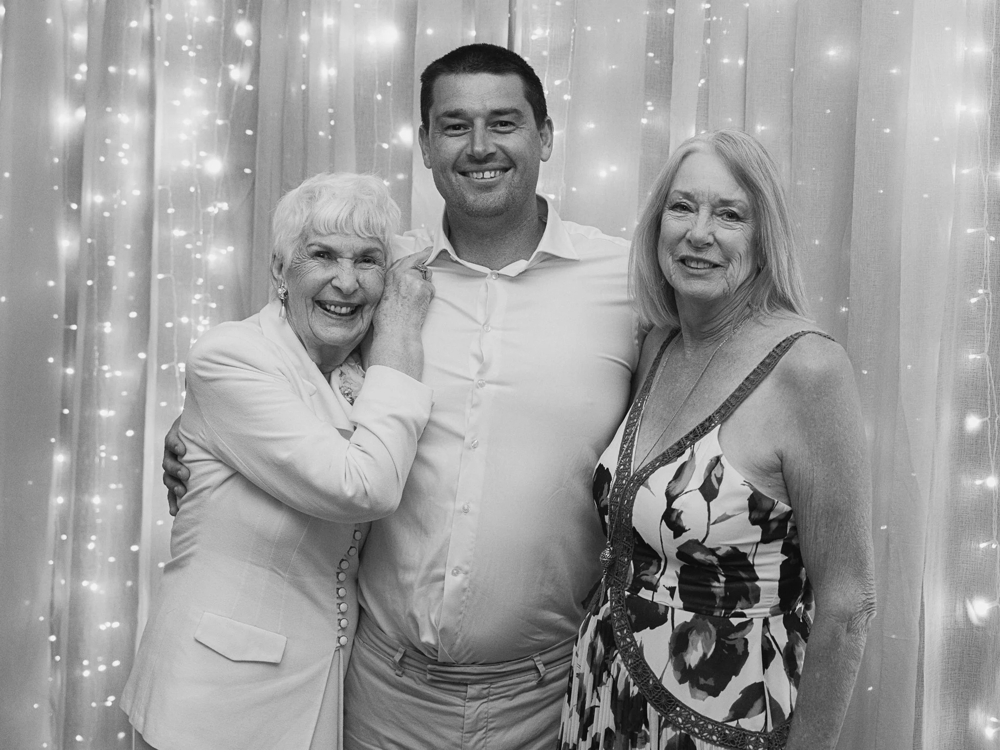 Three people, two women and a man, smiling and posing together in front of a string light background. The woman on the left has white hair and is wearing a light-colored blazer. The man in the middle has short dark hair and is wearing a white dress shirt. The woman on the right has shoulder-length blonde hair and is wearing a patterned dress.