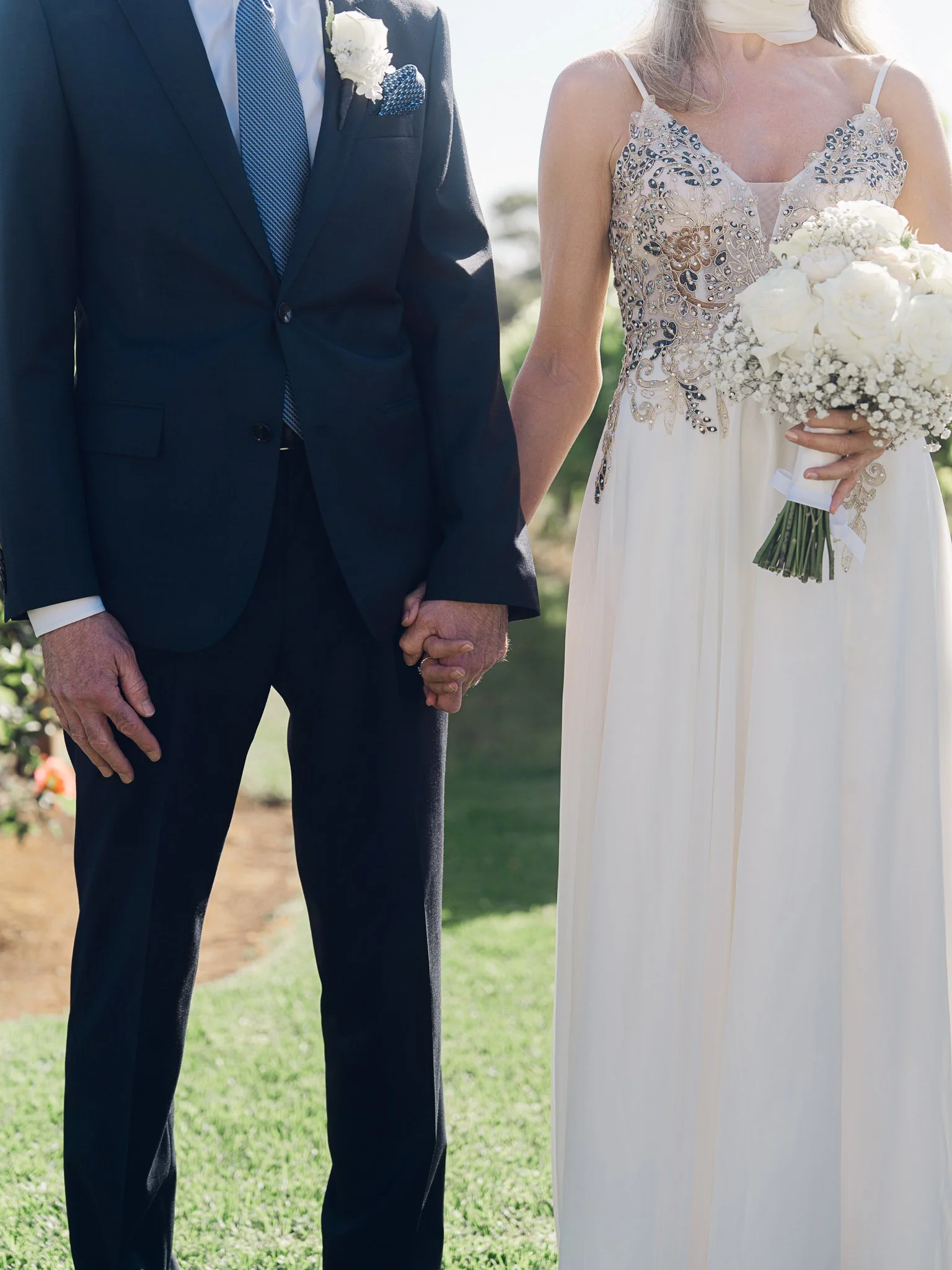 A bride and groom holding hands during their wedding ceremony, with the bride wearing a cream dress with intricate embroidery and holding a bouquet of white flowers, and the groom in a navy suit with a boutonniere and pocket square.