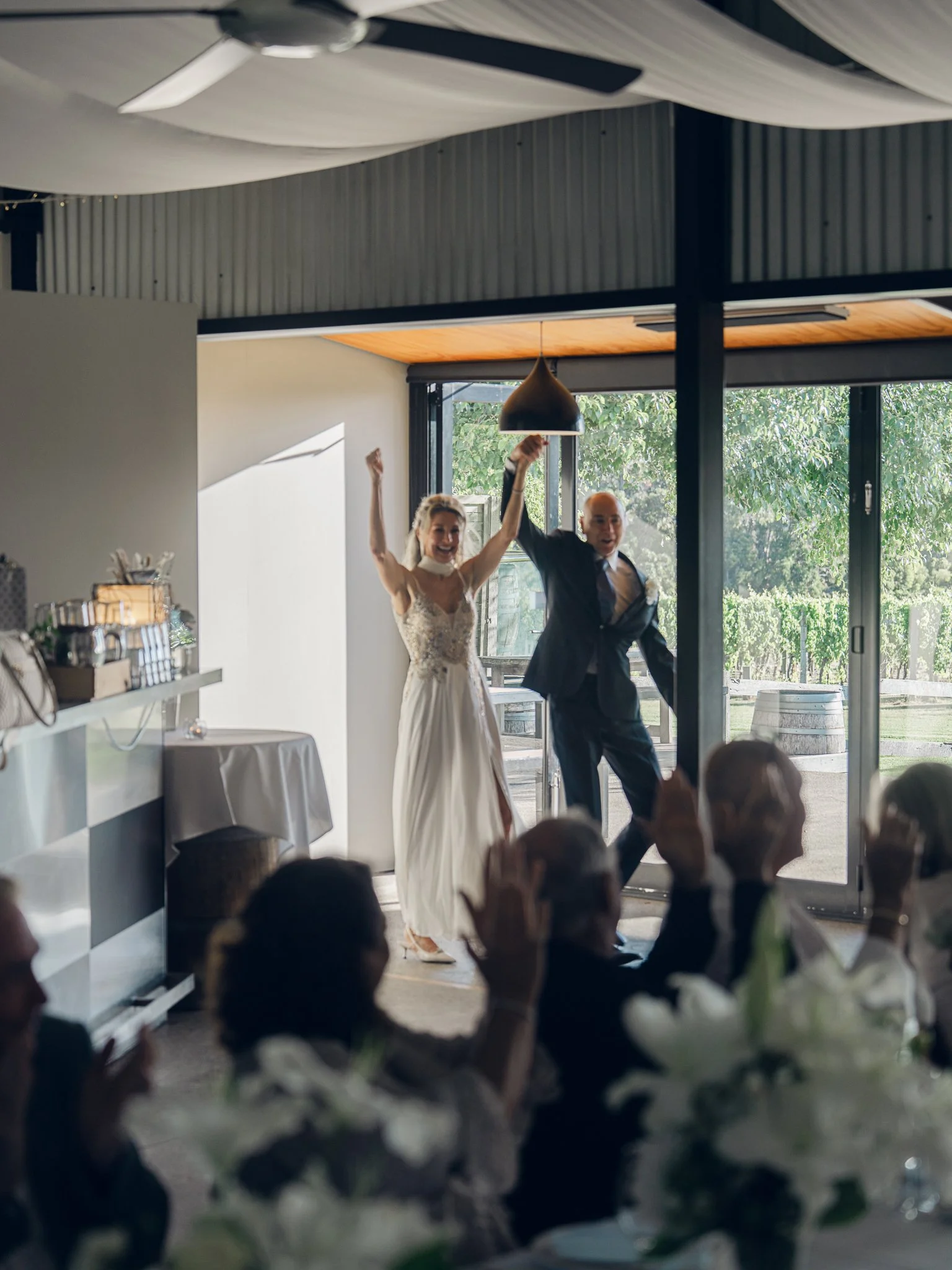 A bride and groom celebrating, holding hands in the air, inside a venue with guests clapping and watching. Bright natural light coming through large glass doors with green outdoor scenery visible.