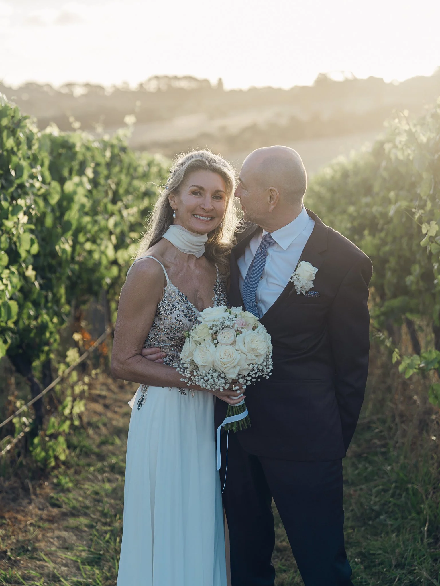 A couple dressed in wedding attire standing together in a vineyard during sunset, with the woman holding a bouquet of white roses and wearing an elegant dress, while the man sports a suit and tie.